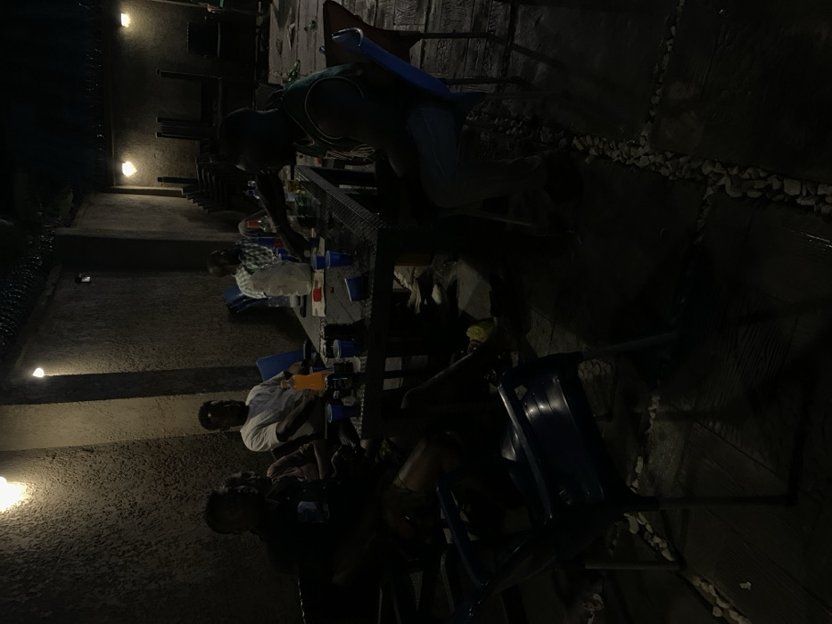 The image depicts a nighttime outdoor gathering of at least six adult males seated around a dark, rectangular table on a patio in Akwanga, Nigeria. The scene is illuminated by multiple wall-mounted light fixtures casting strong shadows.
The individuals are positioned around the table, with four clearly visible figures in the foreground. From left to right: one man in dark clothing, largely obscured by shadow; another in a dark shirt; a third wearing a white t-shirt; and a fourth in a plaid shirt. On the right side of the table, a fifth man wears a green and white sleeveless jersey, facing away from the camera. A sixth individual is partially visible further back on the right.
The table is laden with blue plastic cups, an orange soda bottle, several dark bottles (likely beverages), a white plastic bag or container, and small papers. One man in a plaid shirt is observed reaching towards the table's center.
Seating consists of blue plastic chairs, one of which is empty and prominent in the foreground. In the background to the right, a stack of additional blue plastic chairs is visible. The ground is paved with dark, rectangular tiles interspersed with decorative lines of light-colored pebbles. The surrounding walls are dark and textured, and barbed wire is visible along the top edge of a wall in the upper right.