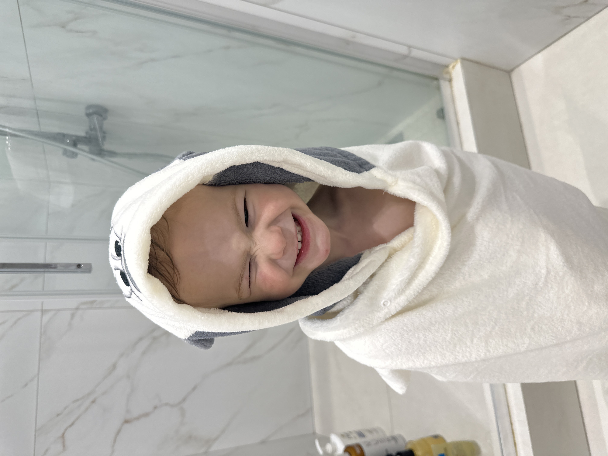 A child is wrapped in a white towel with a gray-trimmed hood, smiling and looking directly at the camera. The setting is a bathroom, with a marble-like wall and a glass shower door visible in the background. There are bottles on a counter. The image is oriented sideways. It is not possible to determine if the image was taken in Прикамский, Russia.