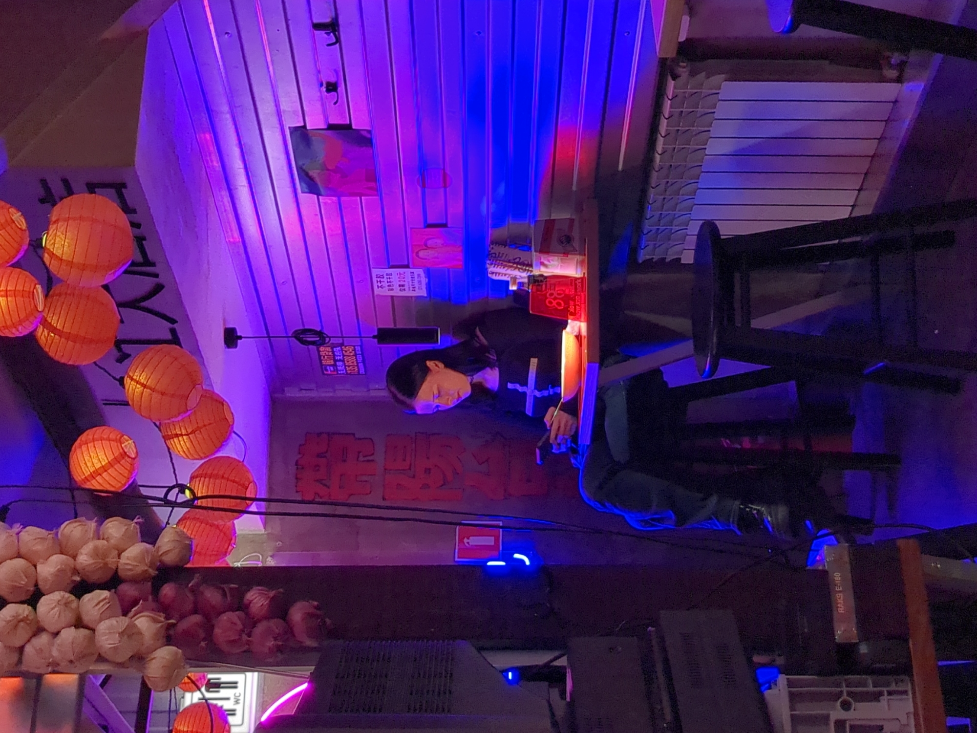 This image captures an indoor scene within an eatery or bar located in Tyumen, Russia. A single young adult female is seated at a small, dark circular wooden table, occupying a dark wooden backless stool. She has dark, shoulder-length hair and is dressed in a dark long-sleeved top, which appears to have a white cross-like design on the chest, dark trousers, and black boots. Her attention is directed towards a smartphone held in both hands, while her right hand simultaneously holds a utensil above an orange bowl containing unidentifiable food. A red plastic cup with visible Chinese characters "慢熟" (mànshú, slow cooking) and "88", along with a clear glass bottle, are also on the table. The establishment's interior is characterized by dim ambient light augmented by vibrant artificial illumination. Multiple orange paper lanterns are suspended from the ceiling, emitting a warm glow. The walls are dramatically lit by blue and purple neon strip lights, particularly a section of horizontally slatted paneling on the right. A pink neon light strip is visible on the far left. Decorative elements include several strings of what appear to be dried garlic bulbs or onions, hanging from the ceiling, with a lower string of red onions or smaller garlic bulbs. The walls display several features: - On the left wall, a red box with a fire extinguisher symbol is mounted, with blue light strips curving below it. A "WC" sign depicting male and female figures is situated further left. A stack of electronic equipment, including a component labeled "RAKS E-180" on a brown box resting on a white crate, is positioned in the foreground on the left. - The back wall features large red Chinese characters, partially obscured, and above the lanterns, additional Chinese characters reading "食入人心" (shí rù rén xīn, food enters people's hearts). - The right wall includes a white sign with black Chinese text indicating "银行换汇" (yínháng huàn huì, bank currency exchange) and phone numbers. Below this, another sign states "不予找零" (bù yǔ zhǎo líng, no change given), "特色干拌" (tèsè gānbàn, specialty dry mix), and "价票 20元" (jiàpiào 20 yuán, ticket price 20 yuan). A framed abstract artwork hangs on this wall, accompanied by two black wall hooks. A white panel radiator is located at the base of the right wall. The floor is dark and reflects some of the blue light. No other individuals or direct interactions are visible in the