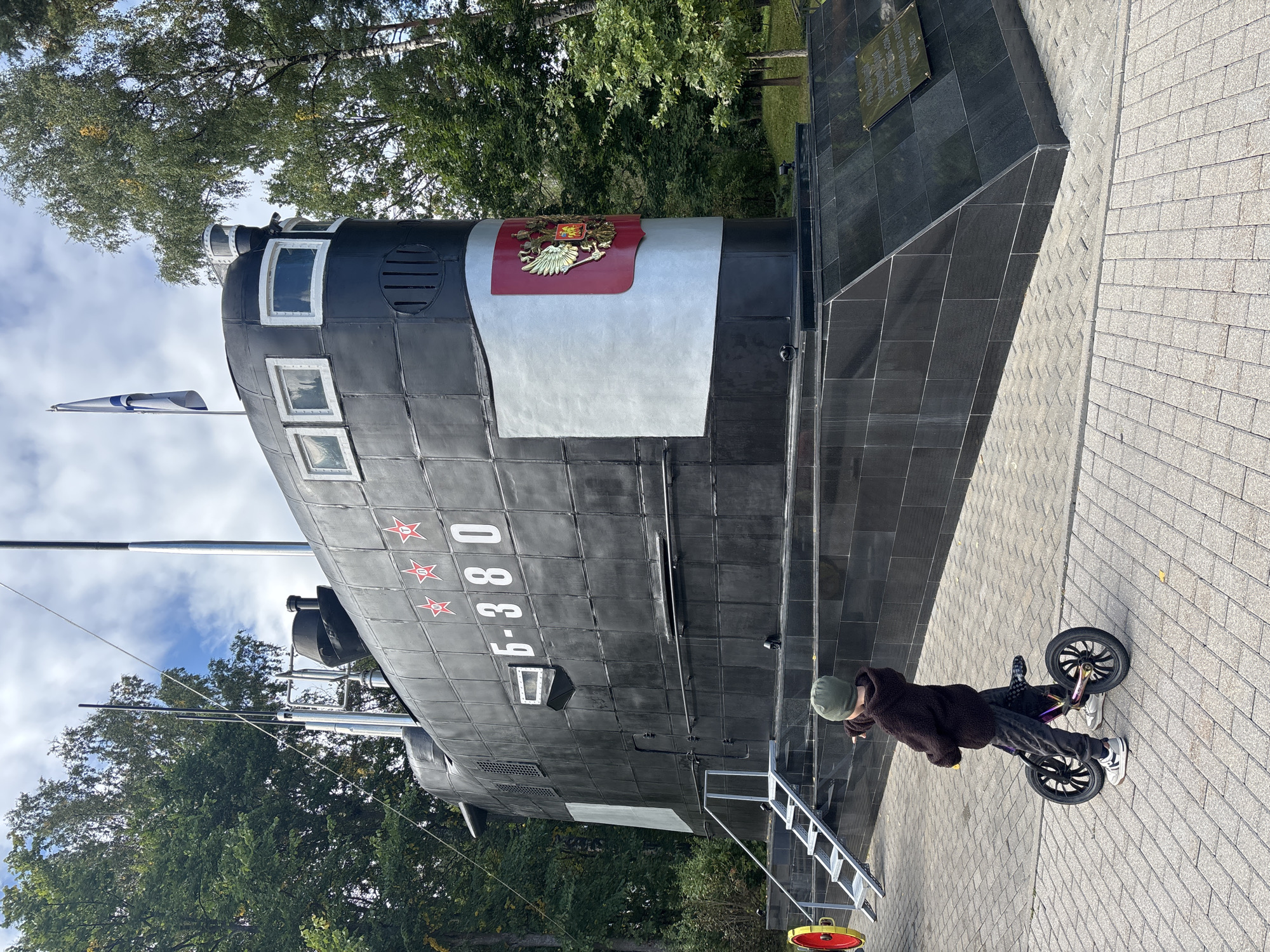 A young child wearing a green beanie, a dark brown hooded sweatshirt, and dark grey pants stands with a balance bike on a paved walkway. The child is facing away from the camera and pointing to the right. To the left of the child, a section of a large, black submarine monument is visible. The submarine has the designation "Б-380" and several red stars on its side. A Russian coat of arms is displayed prominently on the front of the submarine. The submarine sits on a black granite base. In the background, trees and a cloudy sky are present. The scene is located in the city of Рождествено, Russia, likely at a memorial or museum.