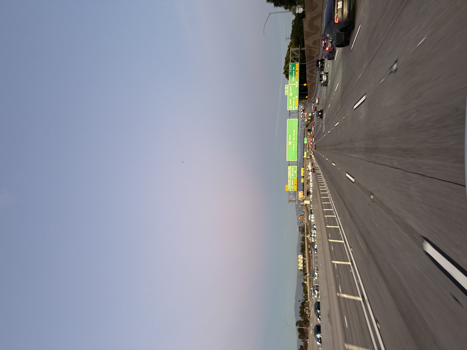A long stretch of a highway with multiple lanes filled with cars during twilight. Overhead green signs are visible, directing traffic towards Irvine, Los Vegas, and San Diego. The sky is transitioning from blue to a soft pink-purple hue, creating a calming backdrop to the congested road. The perspective suggests the photo was taken from inside a vehicle traveling on the highway.