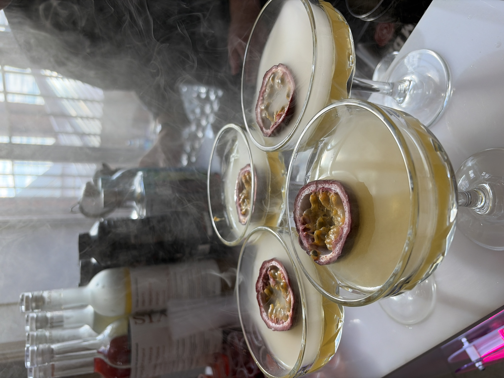 The image shows a close-up, eye-level view of several stemmed glasses containing a light-yellow liquid, each garnished with half a passion fruit. The glasses are arranged on a white surface, likely a bar or table. A rack of liquor bottles can be seen behind the glasses. There is a significant amount of what appears to be vapor or smoke obscuring parts of the scene. The location is unspecified and not identifiable as Neftekamsk, Russia, or any particular city. There is no indication of any people or interactions.