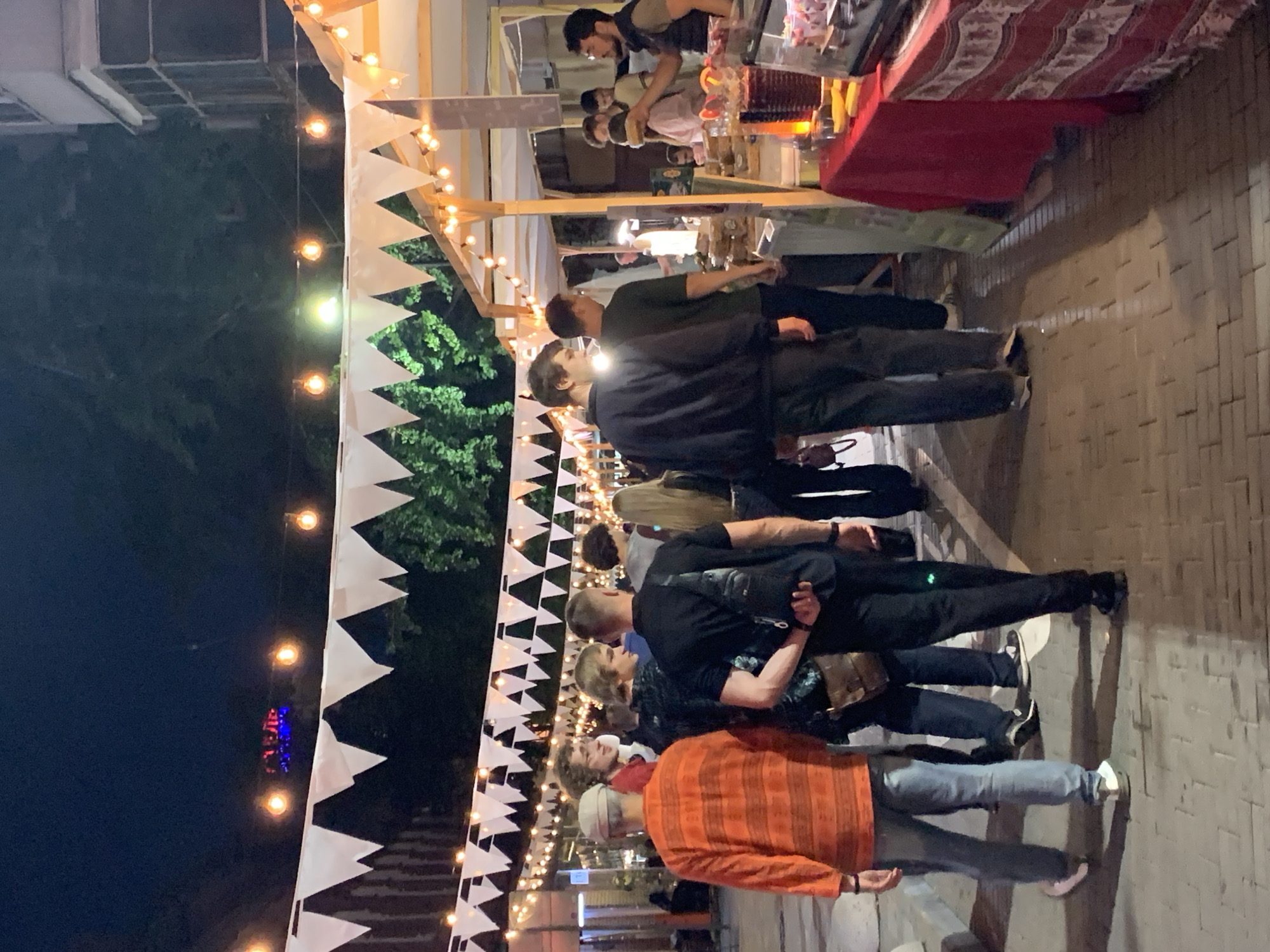 A nighttime street scene shows a pedestrian walkway illuminated by string lights and white triangular flags. Several people are walking along the path, some are browsing market stalls. One stall on the right displays food items on a red tablecloth. The ground is paved with cobblestones. The background features trees and buildings.