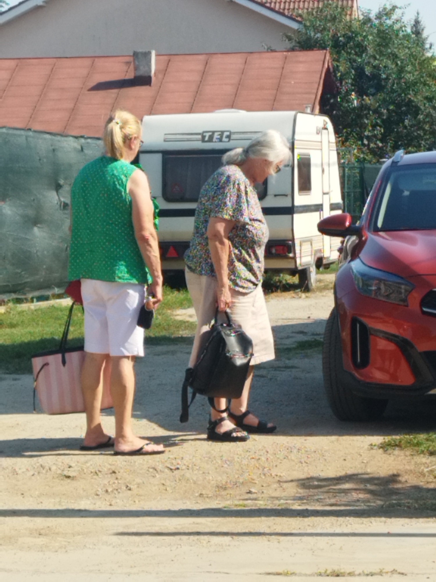 The image shows two women standing outdoors next to a red car and a trailer. The woman on the left is wearing a green sleeveless top with a polka dot pattern, white shorts, and flip-flops. She is carrying a large pink and white striped tote bag. The woman on the right is wearing a floral print shirt and light-colored shorts. She is wearing sandals and carrying a black handbag. The background includes a house with a reddish-brown roof, a green fence, and trees. The scene appears to be on a dirt or gravel surface. The specific location in Bucharest, Romania is not discernible from the image.