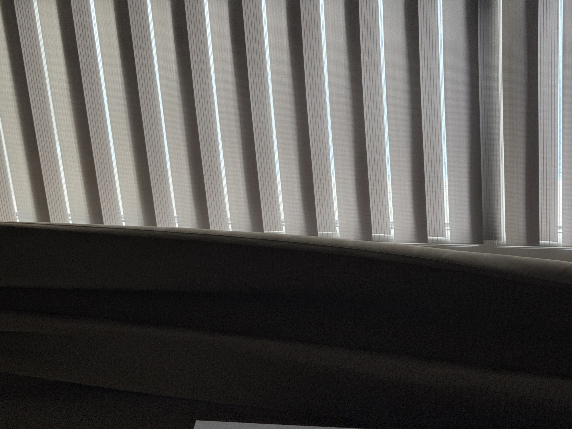 The image displays a close-up of a window with sheer blinds partially covered by a dark curtain. The blinds are made of alternating opaque and sheer horizontal bands, creating a striped pattern. Through the sheer sections, faint daylight is visible, suggesting it is daytime. The curtain on the left side of the frame is a solid, dark color, obscuring a portion of the window. The scene appears to be an interior setting, possibly a home or office. There are no people or discernible actions in the image. The photograph does not provide any visual cues to determine its specific location within Bratislava, Slovakia.
