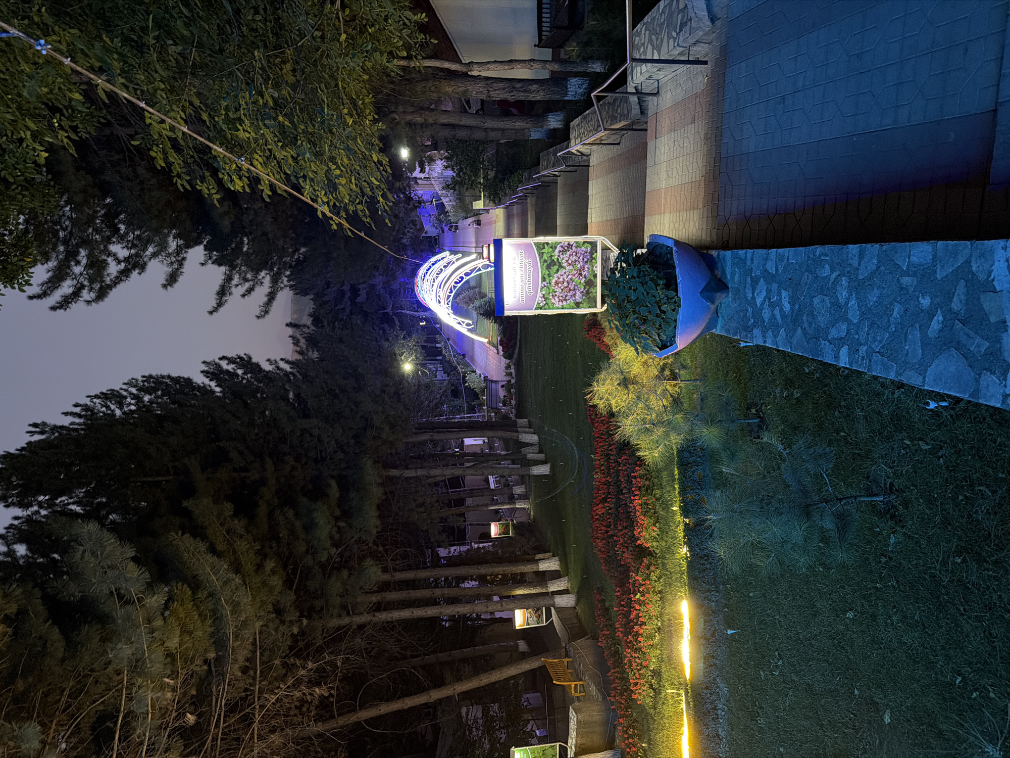 The image depicts a park scene at dusk in Khumsan, Uzbekistan. A paved walkway with stairs ascends from the lower left of the frame towards the upper right, bordered by a stone wall and railings. The walkway is illuminated by decorative blue and white arches that span its width. To the left of the walkway, a manicured lawn is dotted with small pine trees and edged by a vibrant bed of red flowers, illuminated by warm yellow lights. Several tall, slender trees with white-painted trunks line the left side of the lawn, suggesting a planned landscape. A wooden bench is visible on a raised stone platform. Further up the walkway, a large potted plant sits atop the stone wall, and a brightly lit sign with an image of purple flowers and text in an unfamiliar script is mounted on the right side of the path. The sky above is a deep twilight blue. In the distance, beyond the arches, indistinct figures can be seen walking along the path. The overall impression is one of a well-maintained public park designed for evening enjoyment.