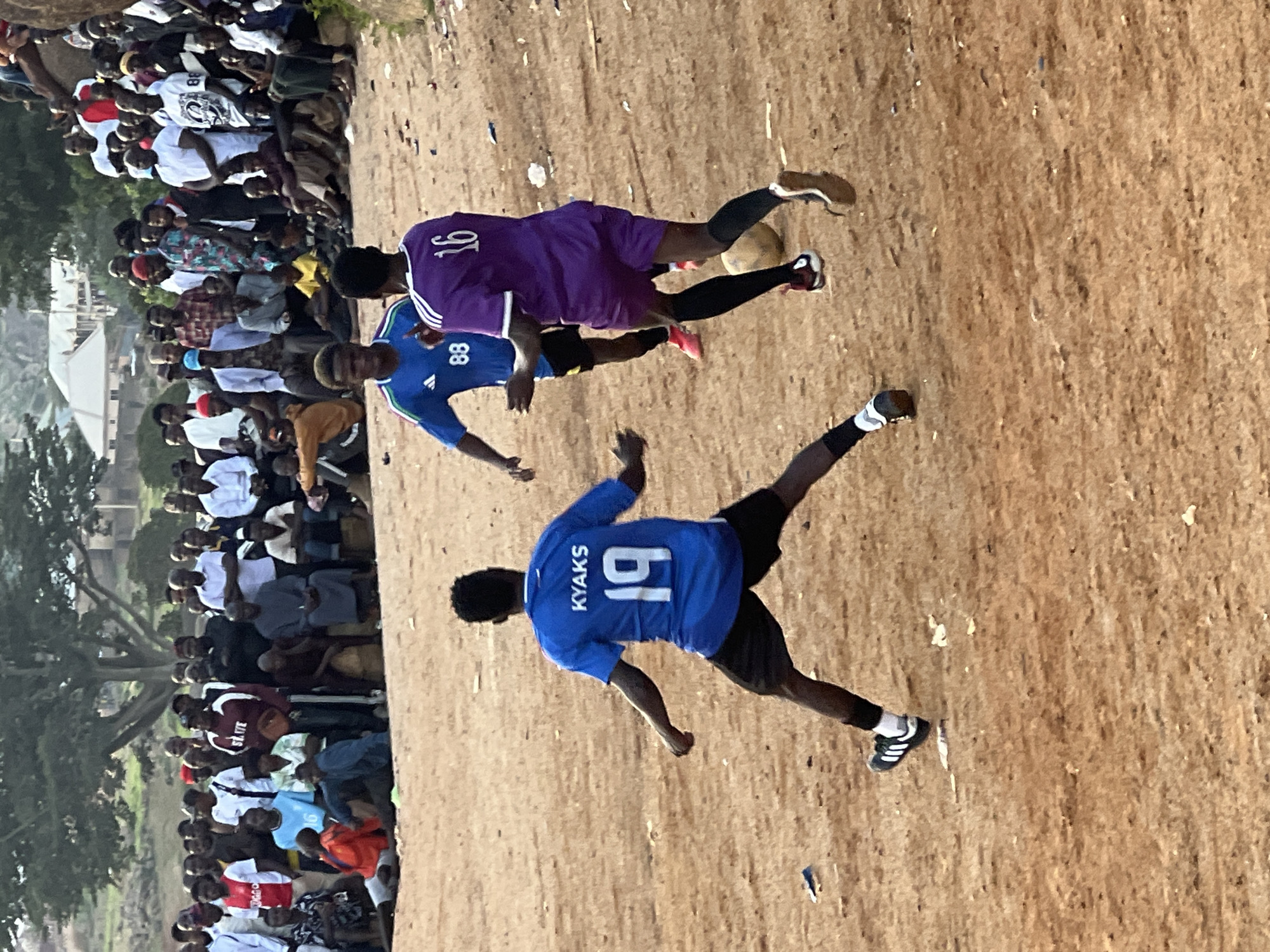 The image captures an outdoor soccer match on a dirt field, likely in Jos, Nigeria. A crowd of spectators lines the edge of the field, some seated and others standing, watching the game. The ground is dusty and uneven, with scattered debris.
Two players are prominently featured in action. One player, wearing a purple jersey with the number "16" on the back, is in motion, possibly kicking or receiving a pass. Another player, dressed in a blue jersey with "KYAKS" and the number "19" visible, is also actively engaged in the game, his body angled as if running or preparing to make a play. Both players are wearing shorts and soccer cleats.
The crowd appears to be diverse, with individuals of varying ages. They are dressed in casual clothing, with some wearing white shirts, suggesting a warm day. The overall atmosphere seems to be one of excitement and engagement, as people are focused on the game.
The background shows some hints of the location. To the left, there are trees and some buildings, possibly indicating the edge of a town or village. The lighting suggests it is daytime, and the overcast sky might imply a possibility of rain or a generally humid climate. The scene depicts a community event centered around a popular sport.