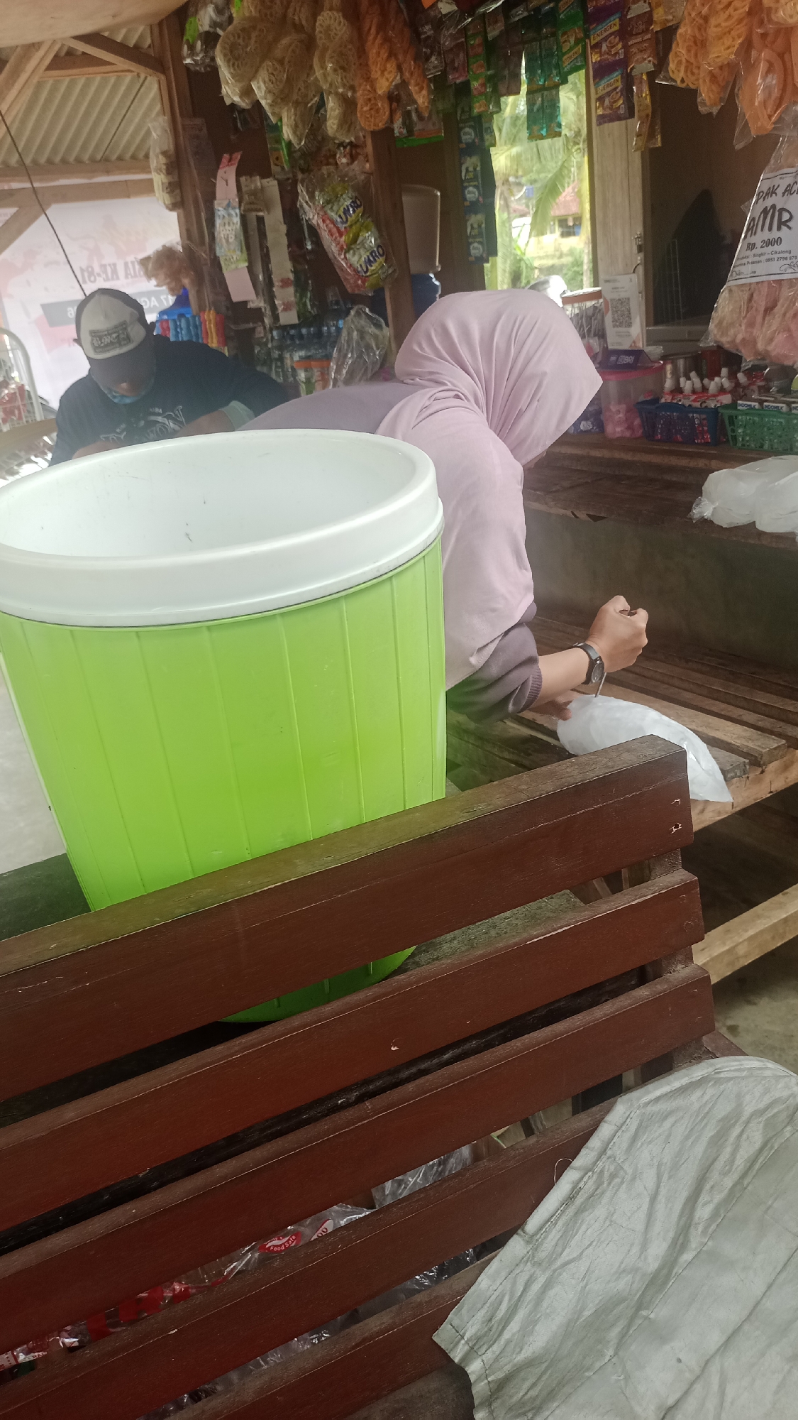 The image depicts a small, open-air convenience store, likely in Cibeber, Indonesia. A person wearing a light pink hijab and a dark grey long-sleeved shirt is seated on a wooden bench, facing away from the camera. They are holding a white plastic bag in their right hand, appearing to be putting something inside it. To their left, another person wearing a black t-shirt and a baseball cap is partially visible, leaning forward.
In the foreground, a large, bright green plastic cooler with a white lid dominates the lower left of the frame. A dark brown wooden bench runs horizontally across the middle ground. Behind the seated individual, shelves are stocked with various packaged goods, including snacks, drinks, and other convenience items. The background shows greenery and hints of a residential structure, suggesting a rural or semi-urban setting. The lighting suggests it is daytime, possibly overcast or shaded.