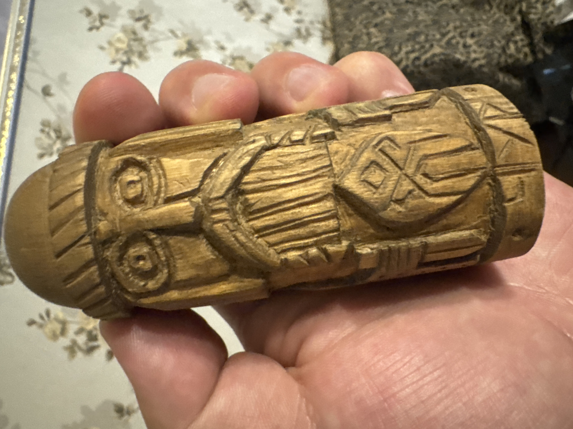 A close-up shot shows a person's hand holding a small, intricately carved wooden object. The object is cylindrical with a rounded end and features detailed carvings that appear to be symbolic or representational. There are circular motifs, geometric patterns, and textured lines carved into the wood. The wood itself has a warm, golden-brown hue. The hand holding the object consists of several fingers, with visible fingernails. The background is out of focus but appears to be an indoor setting with a patterned fabric visible, suggesting a domestic environment. The lighting is soft, implying it might be daytime or indoor lighting. There is no visible text in the image. The focus is entirely on the hand and the carved wooden object, highlighting the craftsmanship of the carving.