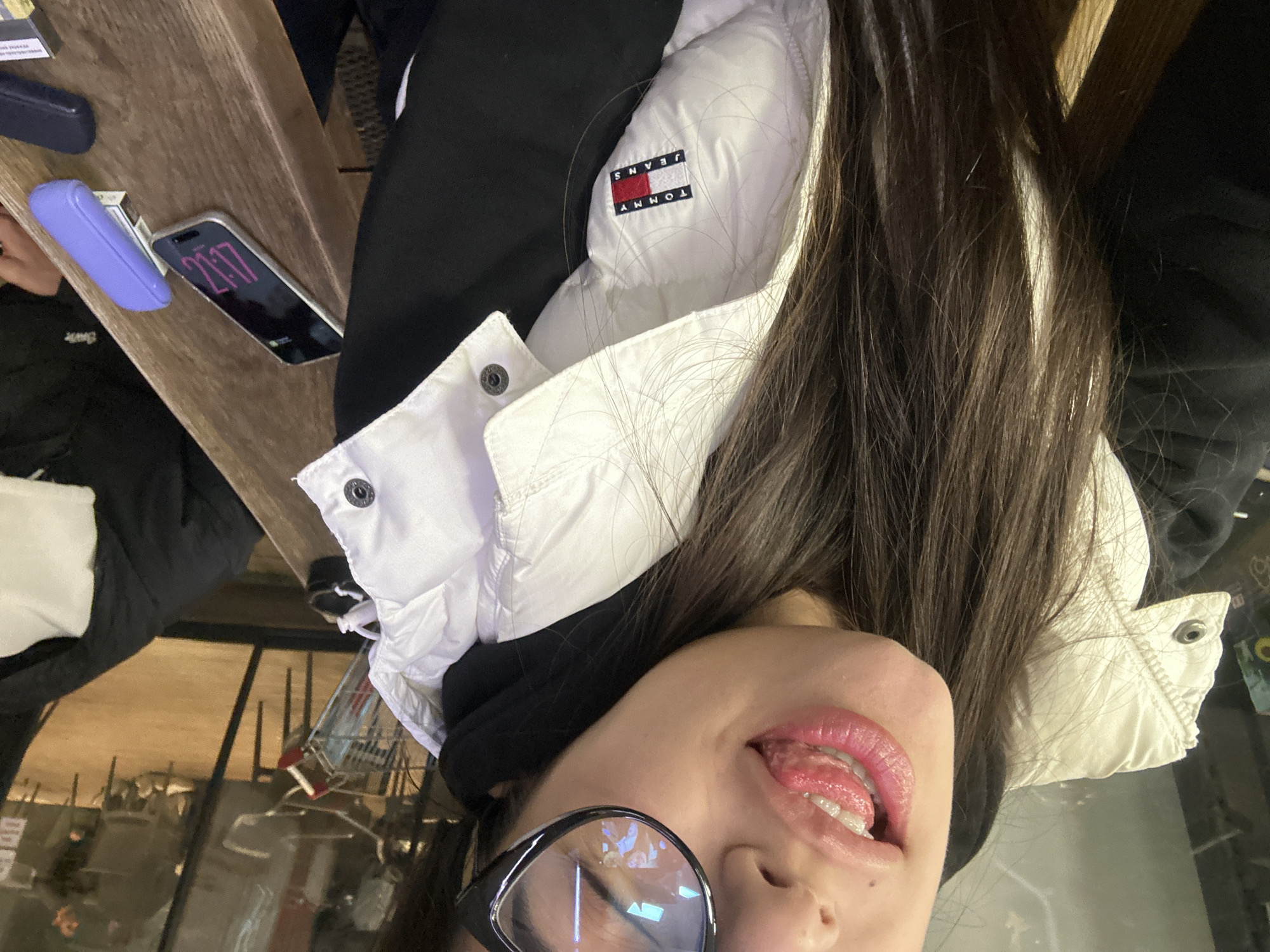 A rotated, close-up shot shows a person with long, dark hair and glasses. The person is wearing a white and black Tommy Jeans jacket. The person's tongue is visible. To the left, on a wooden surface, are a phone displaying "21:17," a blue rectangular object, and what appears to be a cigarette pack. The background shows a blurry reflection of the interior of a restaurant or cafe. Considering the request for location, and based on the visual appearance of the interior, it's plausible the image was taken in a cafe or restaurant in Plovdiv, Bulgaria.