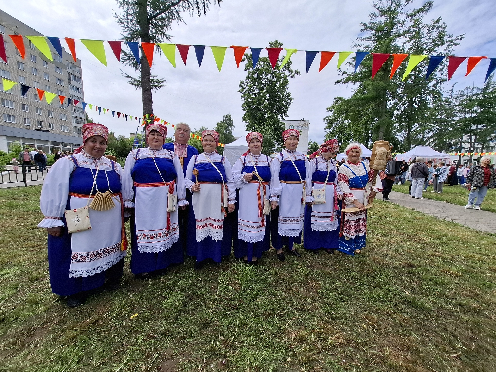 An outdoor event is captured in Chaikovsky, Russia, with a group of eight individuals in traditional clothing standing in a line on a grassy area. The individuals, appearing to be participants or performers, are adorned in blue and white embroidered attire with red headscarves. They are positioned in front of a multi-story building and a line of colorful triangular flags strung between trees. The background reveals more trees, tents, and a paved walkway with other people in casual attire. The weather appears overcast, contributing to the diffused natural light of the scene.