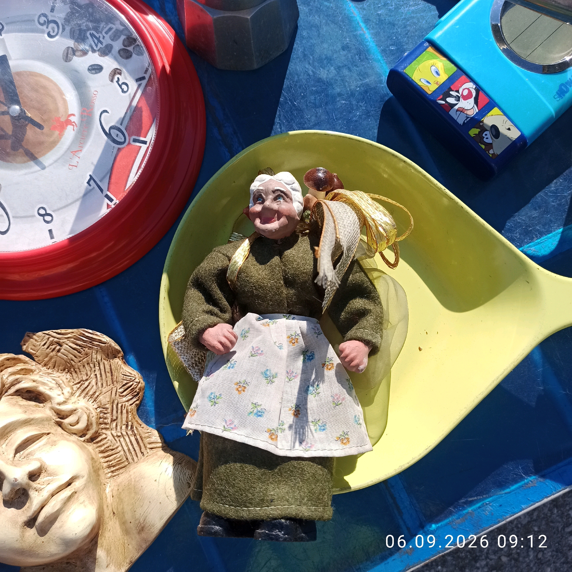 On a blue tarp, a close-up shot captures a scene of various items. In the foreground, a carved relief of a face, rendered in a light beige material, is positioned on the left. To its right, a doll depicting an elderly woman with white hair, rosy cheeks, and blue eyes is seated within a yellow dish-shaped object. The doll wears a dark green long-sleeved top and a white apron with a floral pattern. A decorative ribbon, metallic gold and white, is draped around the doll. Further to the right, a blue electronic device with cartoon characters, possibly a portable game or music player, is visible. In the upper left quadrant, a red alarm clock with a white face and Roman numerals is partially visible, with coffee beans scattered on its surface. Behind these items, a dark grey, hexagonal object, possibly a decorative stone or a piece of hardware, is present. The overall environment appears to be an outdoor setting, with the presence of shadows suggesting daylight. A timestamp "06.09.2026 09:12" is overlaid on the bottom right of the image.