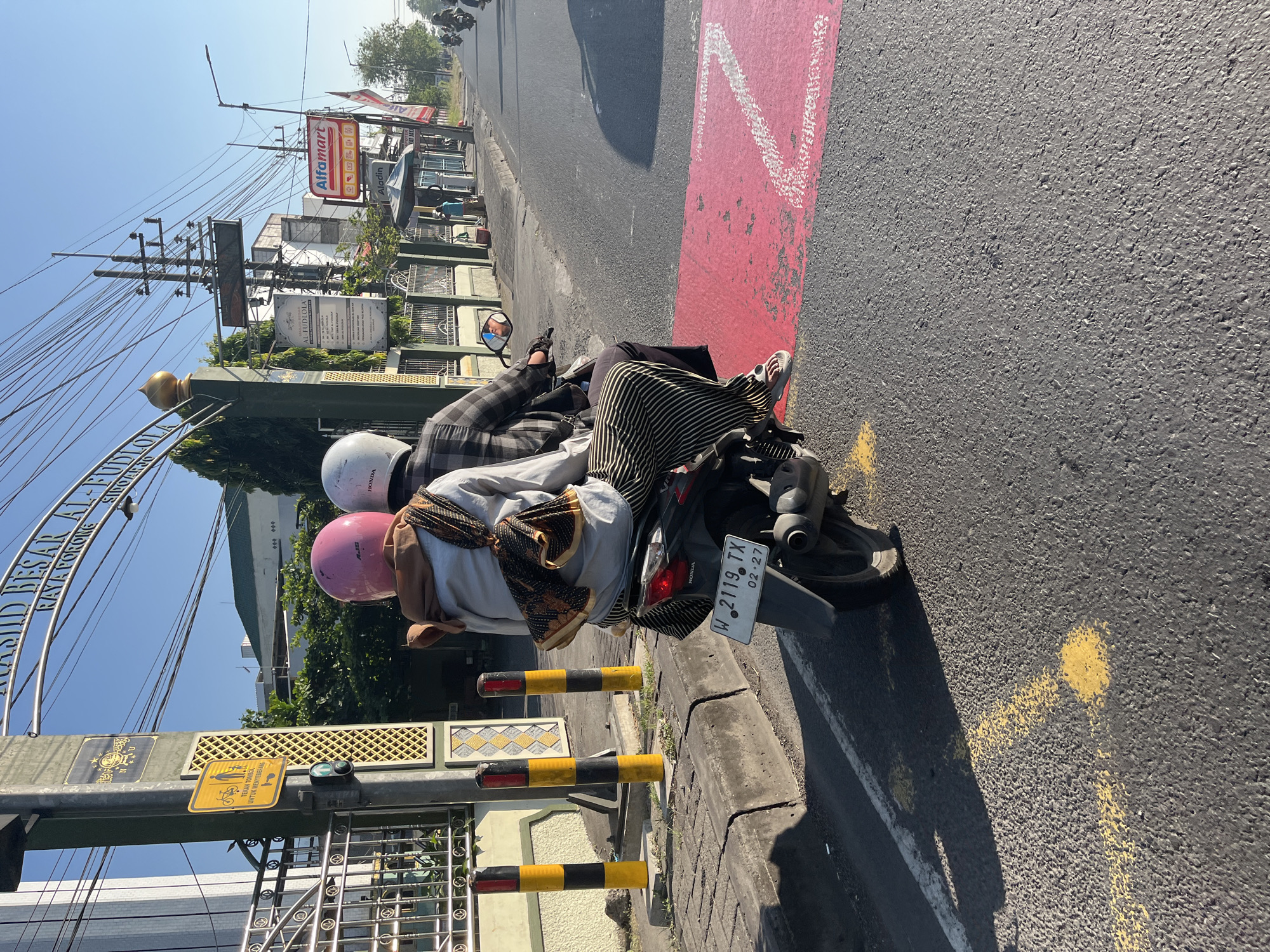 Two individuals, an adult and a child, are seated on a motorcycle, viewed from behind. The adult, in the front, wears a white helmet and a plaid shirt, while the child behind wears a pink helmet and a brown scarf. The child is also wearing a striped skirt. The motorcycle is stopped at a crosswalk marked with red paint and yellow markings on the road. In the background, there is a building with a sign that reads "MASJID BESAR AL-FUDLOLA" and "RAYA PORONG SIDOARJO". There are also utility poles with many wires, a sign for "Alfamart", and other buildings and traffic visible on the street. The scene appears to be in a city with moderate traffic and a clear blue sky.
