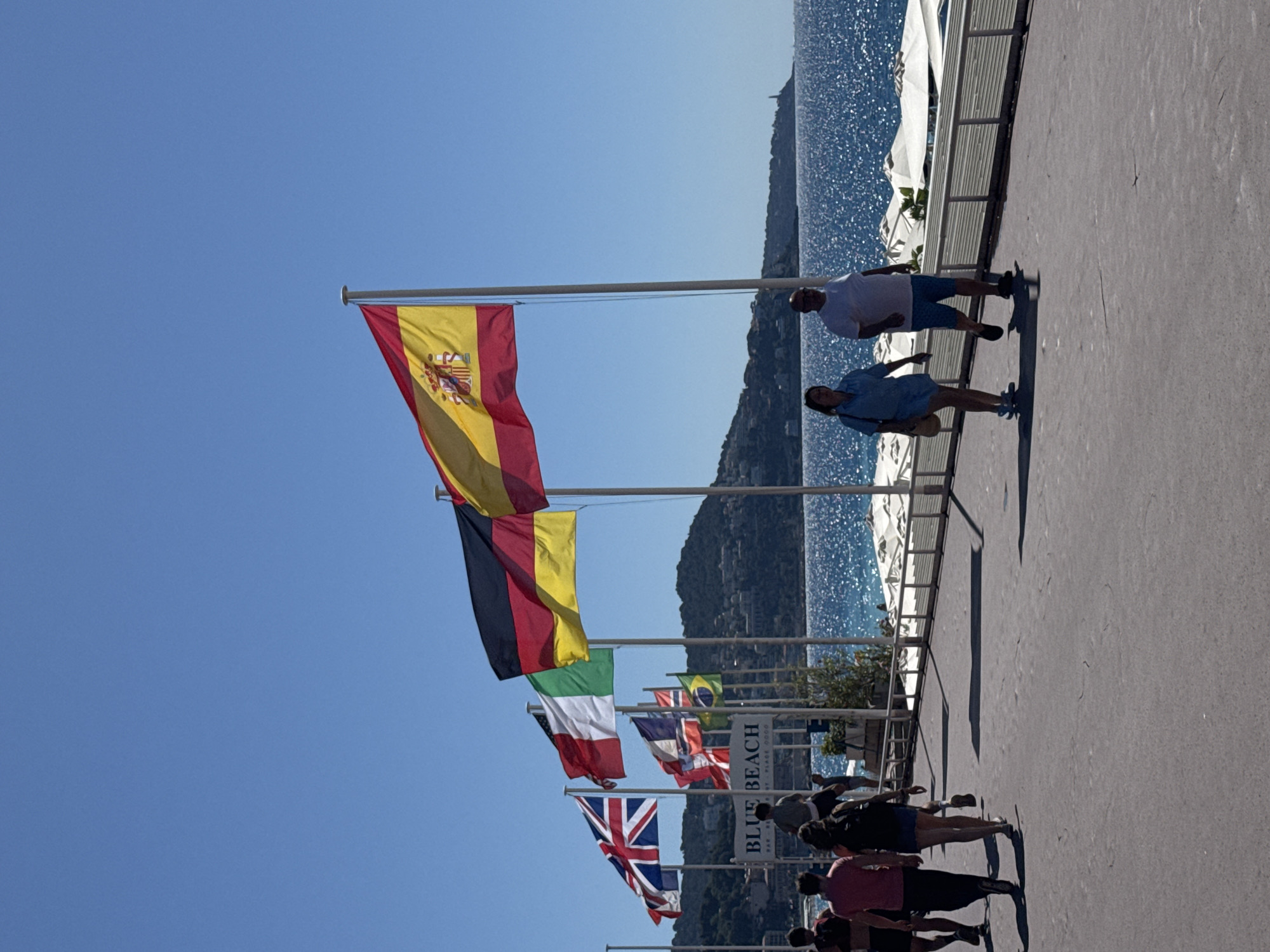 The image depicts a sunny day at a seaside promenade in Nice, France. Several national flags, including those of the United Kingdom, Italy, Germany, and Spain, are prominently displayed on flagpoles along the walkway. In the background, the Mediterranean Sea sparkles under the bright sun, with a coastline featuring buildings and a lighthouse visible in the distance. A sign indicating "BLUE BEACH" is present, suggesting this is a beach club or resort area. Several people are walking along the promenade, some dressed casually for a warm day. Two individuals in the foreground are rollerblading. The overall atmosphere is relaxed and summery.