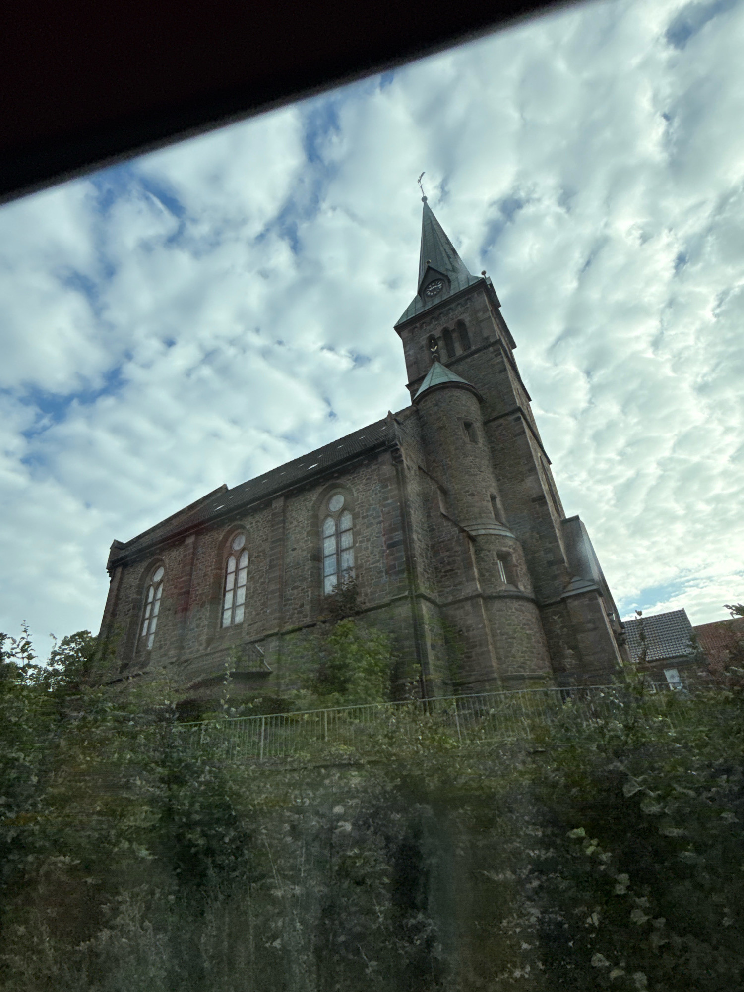 The image shows a brick church with a tall steeple against a cloudy sky. The church has arched windows and a clock on the steeple. It is surrounded by trees and greenery. In the background, there are some houses with tiled roofs. The image is taken from a low angle, looking up at the church. The location is identified as Herzberg, Germany.