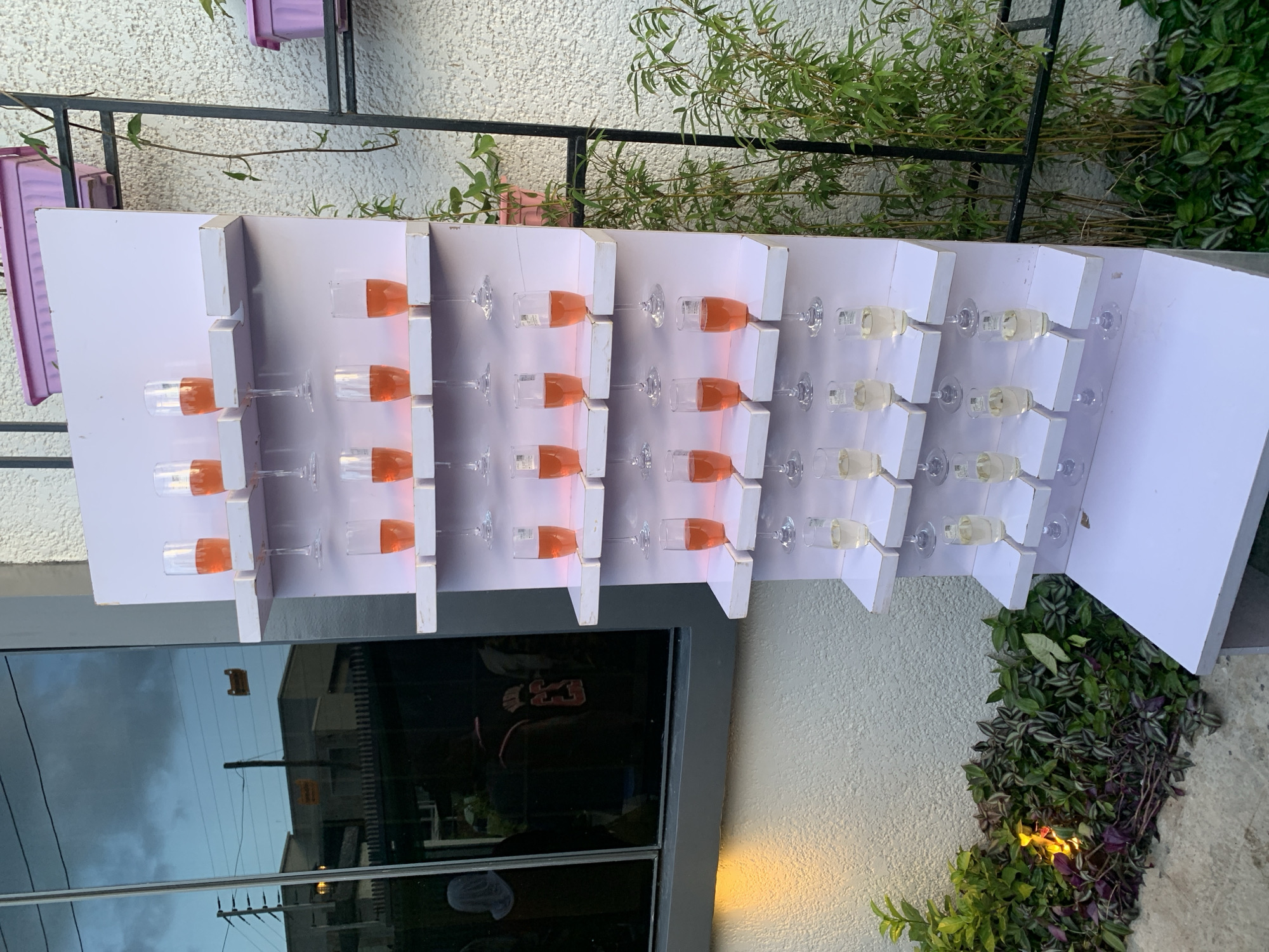 The image shows a white, tiered display stand filled with champagne flutes. The flutes on the upper levels contain an orange liquid, likely champagne or a similar beverage, while those on the lower levels appear to contain a clear, pale yellow liquid. The stand is positioned outdoors, with a textured wall and lush green foliage to the right. To the left, a large window reflects the sky and buildings, suggesting an urban or semi-urban setting. The overall scene suggests a celebratory event or catering setup. The location is specified as Gura Top, Nigeria. There are no discernible people in the immediate vicinity of the display stand.