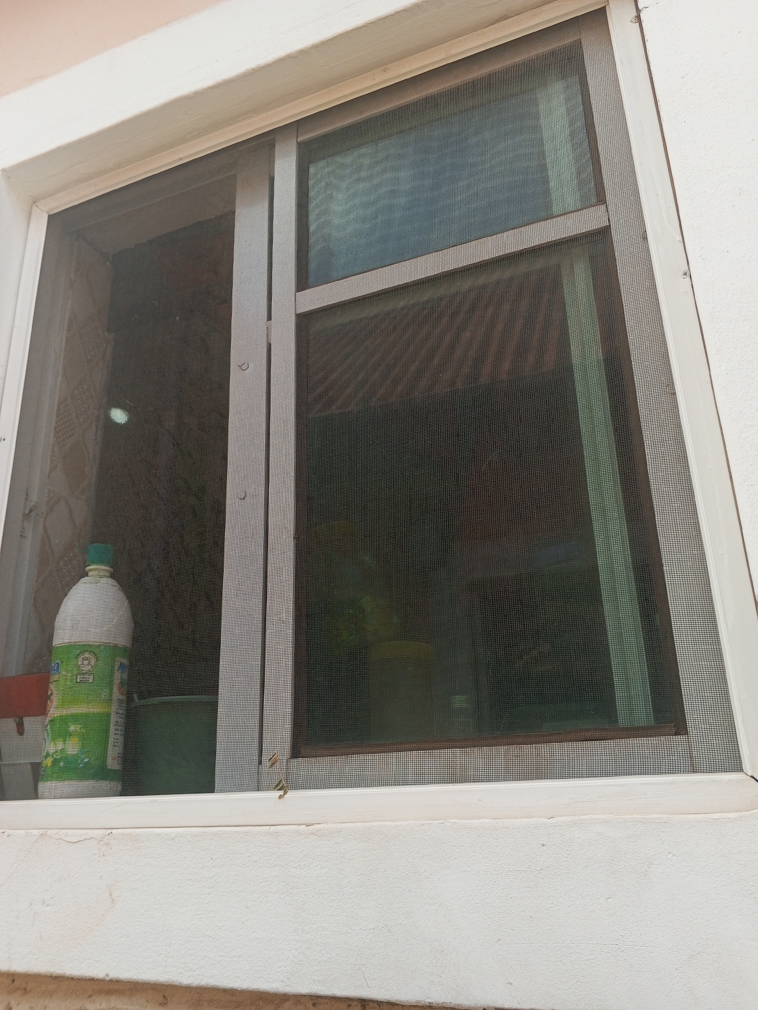 The image shows a window with a white frame and a mosquito screen. A bottle and a green container are visible on the windowsill. The view through the window is obscured by the screen. The context of Abuja, Nigeria, is not discernible from the image alone; this cannot be confirmed from the visual data.
