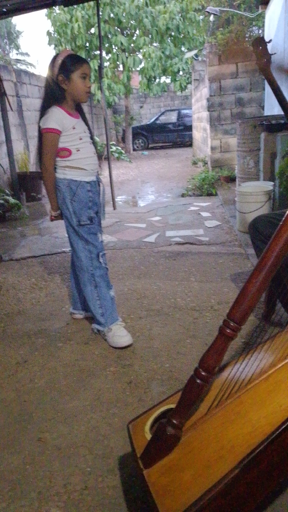The image shows a young girl in Tinaquillo, Venezuela. She is standing in a courtyard or backyard, wearing a white t-shirt with pink trim, blue jeans, and white sneakers. A black car is parked in the background, visible through an opening in a wall. In the foreground, there is a Paraguayan harp. The courtyard is paved and has a few plants around. There is a white bucket near the harp. A brick wall is visible on the right side of the image.