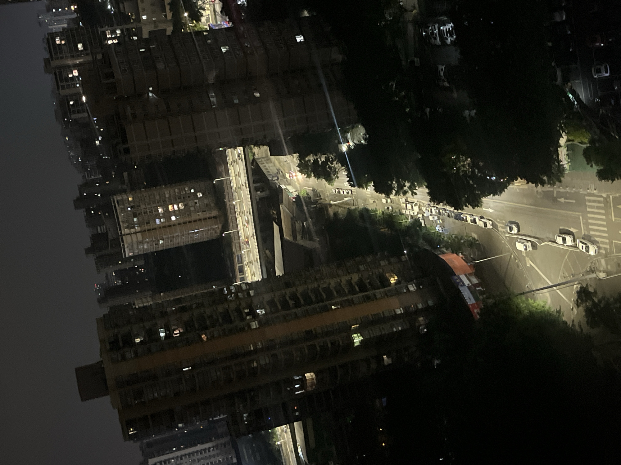 This image depicts a nighttime urban landscape, identified as Jiulongpo District, China, captured from an elevated perspective looking down and across the scene at an oblique angle. The environment is characterized by numerous multi-story residential or commercial buildings, many with illuminated windows, indicating occupancy.
A primary street traverses the mid-ground, running diagonally from the lower left to the upper right, illuminated by streetlights. Multiple vehicles, predominantly white passenger cars, are visible parked along this street and in an adjacent parking area to the right. A white arrow on the asphalt indicates a one-way traffic flow. In the background, an elevated multi-lane highway or bridge is visible with moving vehicle lights.
Dense, dark tree canopy flanks sections of the street and forms significant shadows, particularly in the foreground and along the right side. On a building or structure visible in the middle-right area near the street, a banner displaying red, white, and possibly blue/green sections with indiscernible text or logos is present. No individual people are clearly discernible, but the illuminated windows and active streets imply human presence. The overall lighting suggests an urban area operating after dark.