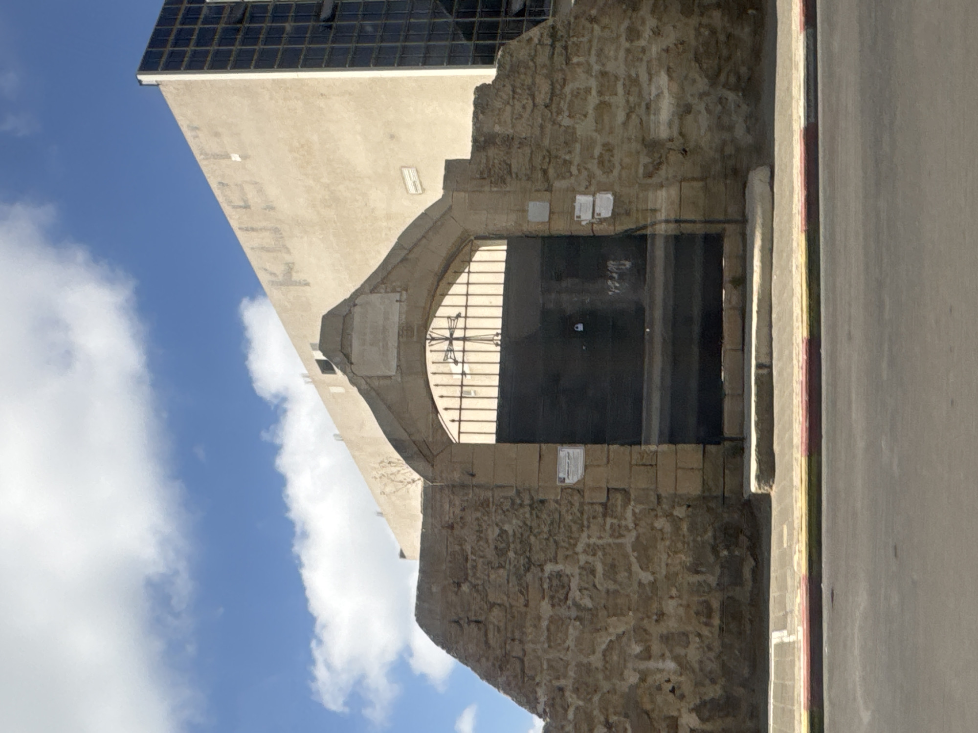 The image shows an arched gateway leading to an entrance with a metal gate, flanked by stone walls and a building in the background. The structure appears old, with textured stonework. The building in the background has a modern aesthetic. The sky with scattered clouds suggests daytime. The scene is located in the city of Tel-Aviv, Israel.