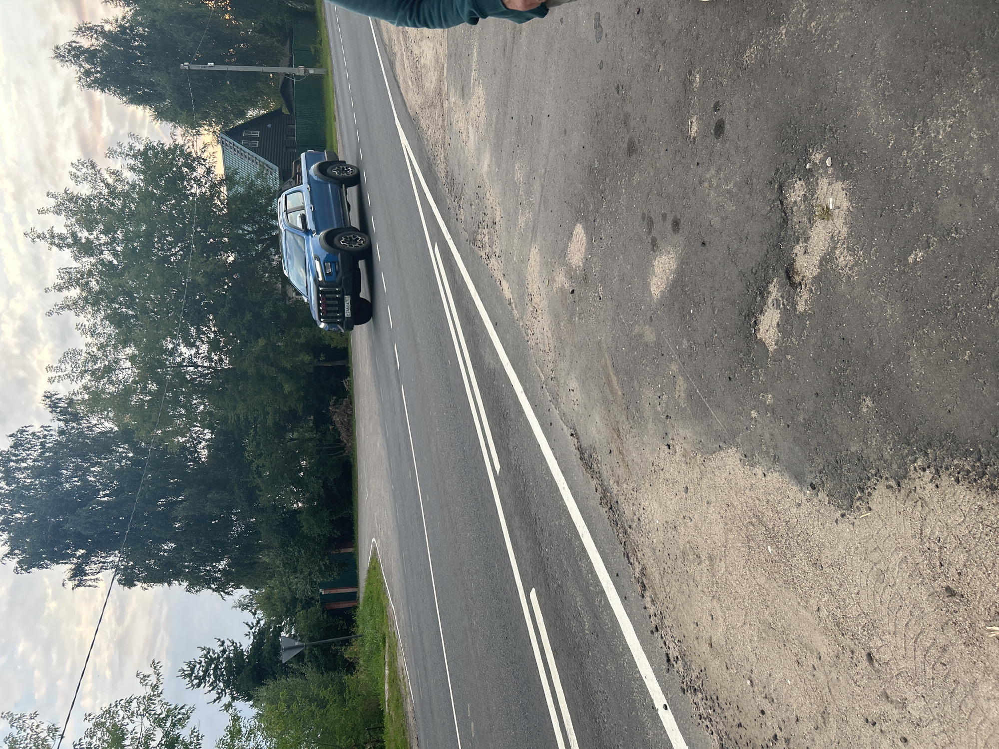 The image depicts a roadside scene in Kalitsino, Russia, with a blue pickup truck driving on an asphalt road. The road has visible white lane markings and some patches of wear and tear, including potholes and areas where the asphalt has crumbled away, revealing sand and dirt. To the left of the road, there is a dirt shoulder with tire tracks visible in the sand. Beyond the road, a dense line of green trees forms a backdrop, with a small wooden house with a dark roof visible through the foliage. A metal gate and a signpost are also present near the tree line. The sky is overcast with clouds, and the lighting suggests it is either early morning or late afternoon. A person's right arm and shoulder, wearing a teal-colored sleeve, are visible in the lower right corner of the frame, indicating the photographer's presence at the scene.