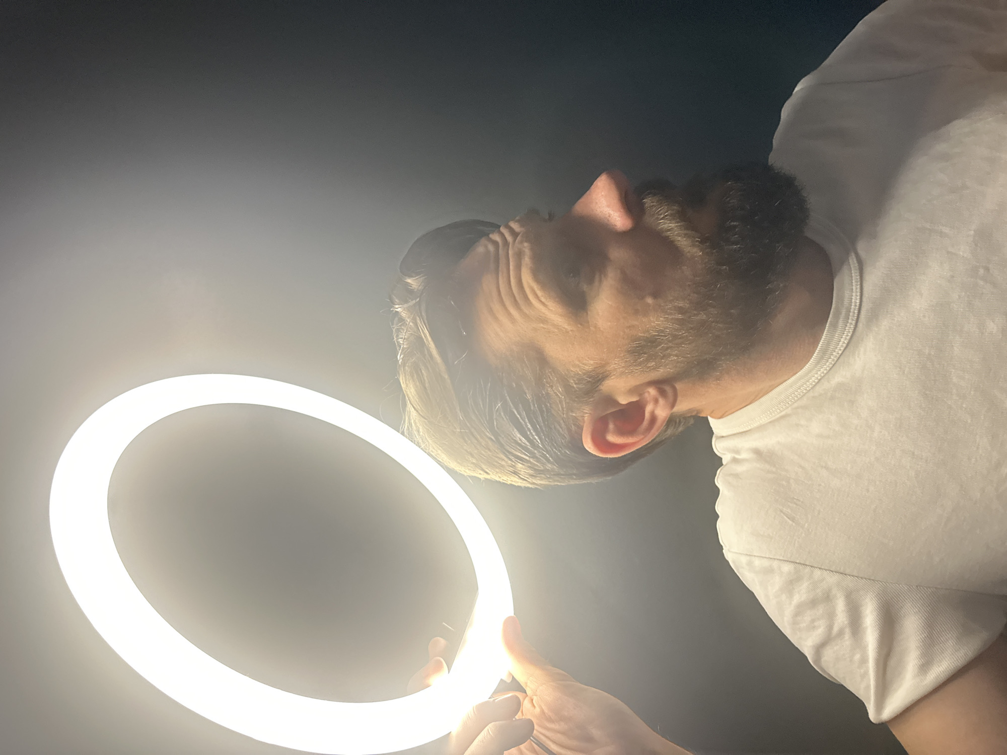 The image captures a close-up, low-angle perspective of a man interacting with a bright ring light. The man, situated in Fethiye, Turkey, is seen from the chest up, wearing a plain white t-shirt. He has short brown hair and a dark beard. His head is sharply tilted downwards and to the left, as if he is looking intently into or past the ring light. His left hand is visible at the bottom of the frame, holding the illuminated circular light.
The ring light, a prominent object, emits a strong, diffused white light, creating a halo effect. The light strongly illuminates the man's face from below, highlighting the contours of his nose and the wrinkles on his forehead, suggesting concentration or a reaction to the bright light. His right ear is also visible, illuminated. The background is dark and indistinct, likely a wall or an unlit room, emphasizing the man and the light source. The overall impression is one of a controlled lighting setup, possibly for photography or videography, with the man adjusting or examining the light. No discernible text is visible in the image.
