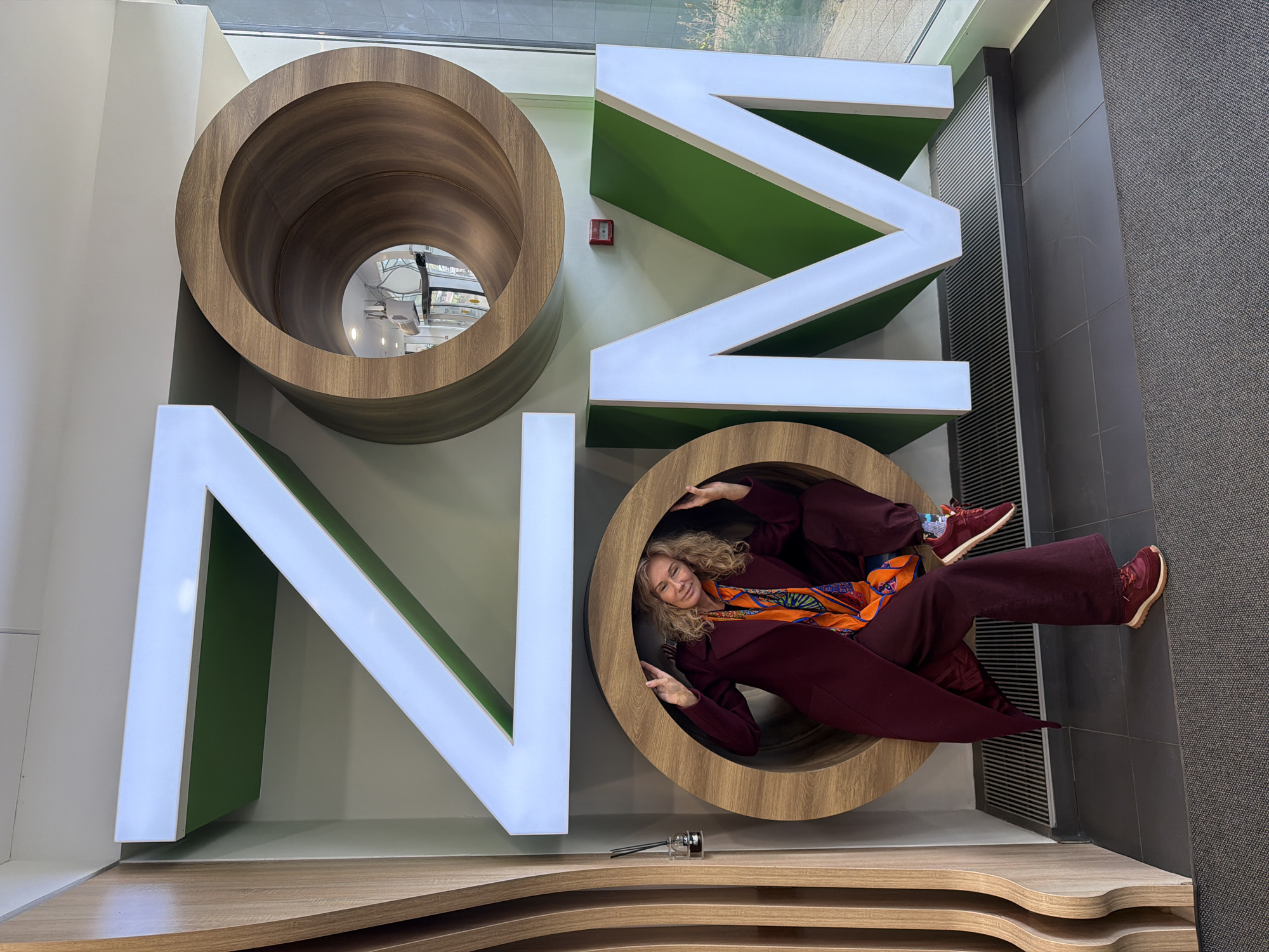 A woman is lying inside the second letter "O" in a large decorative word "ONMO" on a wall, likely in an interior space. The letters appear to be constructed from different materials: "O"s are made of wood, and "N" and "M" have white exteriors and green interiors. The woman is wearing a maroon outfit with a colorful scarf and maroon sneakers. She is smiling and has her hands raised near her face. A small silver object is visible on the shelf in front of the "O." There is also a red alarm-like button above the letter "M". The wall is white, and gray tiles can be seen on the right-hand side. A window reflects an exterior scene. Without more information, the location in Saint Petersburg, Russia cannot be confirmed.