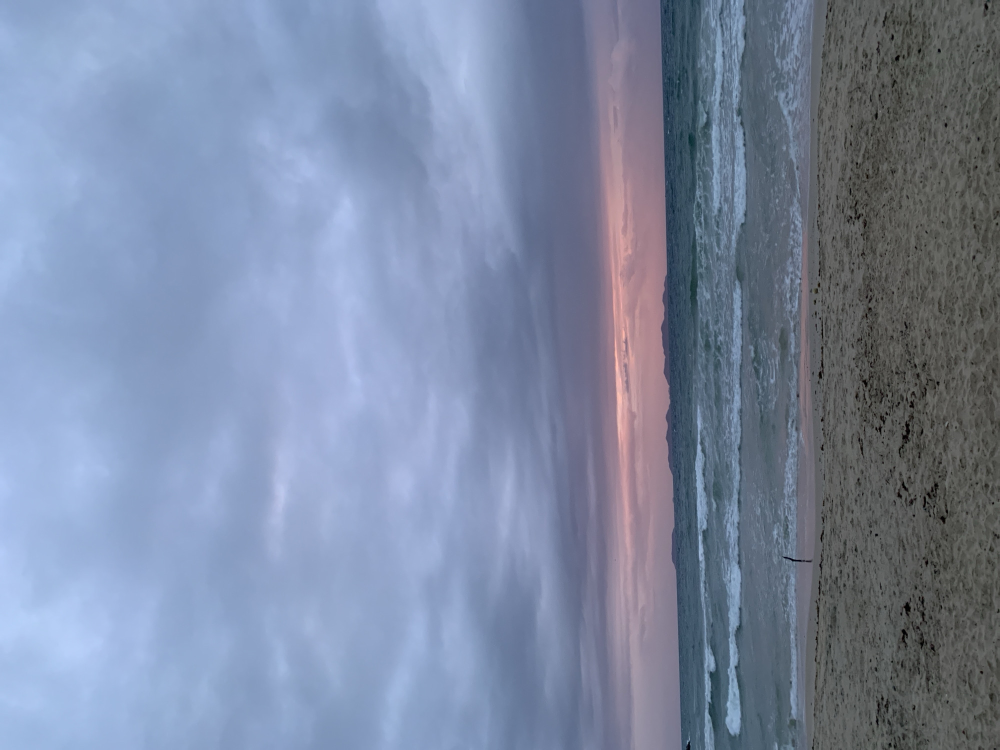 The image depicts a tranquil beach scene at dusk in El Tirano, Venezuela. The sky is filled with dramatic, gray clouds, hinting at an approaching or departing storm, with a subtle blush of pink and purple light visible on the horizon where the sun has set. The sea is a deep teal, with moderate waves crashing onto the shore, creating white foam. In the foreground, a sandy beach stretches across the frame, with a single, dark, stick-like object standing upright in the wet sand near the water's edge. In the distance, a faint, mountainous landmass is visible against the horizon. There are no people or animals in the frame, and the overall impression is one of natural beauty and solitude.
