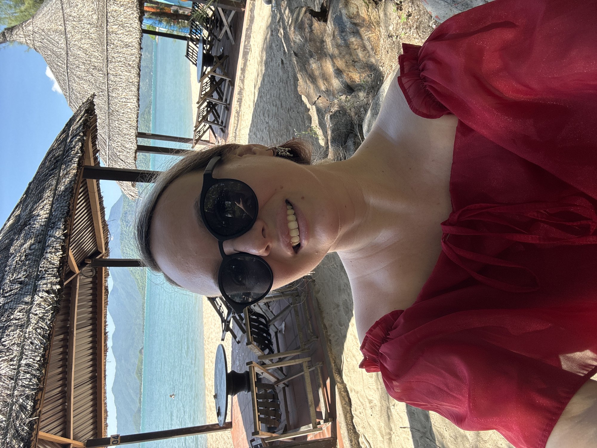 A woman is taking a selfie on a sandy beach with thatched-roof structures and lounge chairs in the background. She is wearing a red off-the-shoulder top and black sunglasses. The ocean and a lush green landscape with mountains are visible behind her. The lighting suggests it is a sunny day.
