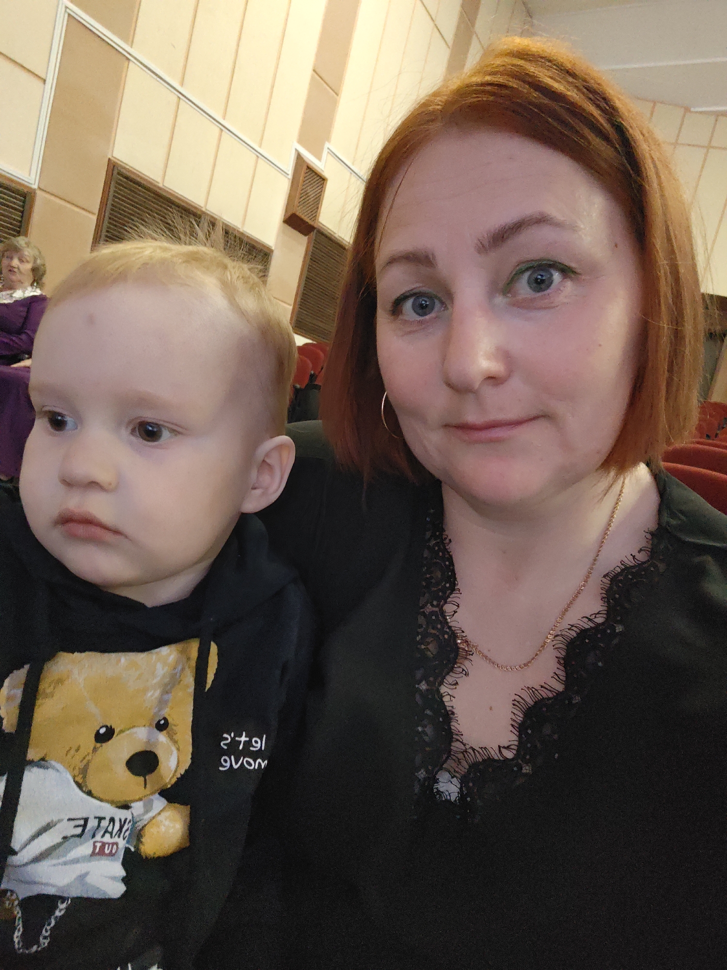 The image shows a selfie of a woman with red hair and a fair-skinned toddler. The woman is wearing a black top with lace detailing around the neckline, gold hoop earrings, and a gold chain necklace. She has blue eyes and appears to be wearing green eyeliner. The toddler, positioned to the left of the woman, is wearing a black hoodie with a teddy bear graphic on it.
In the background, there are panels on a wall, and an elderly woman is partially visible to the left. Red seating can also be seen. Based on the location provided, the image was taken in Марковский, Russia. There are no apparent actions or interactions besides the selfie being taken. The environment suggests they are indoors, possibly in a theater or public venue.