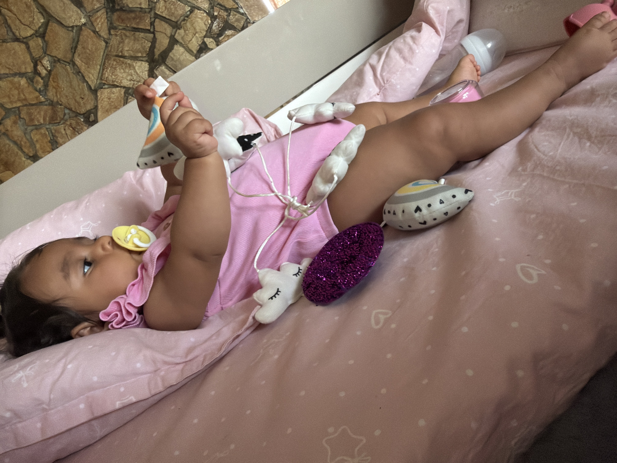 A baby girl is lying on a pink, polka-dotted blanket in an indoor setting. She is wearing a pink dress and has a yellow pacifier in her mouth. A mobile with various plush toys, including clouds, a glittery oval, and a bird-like toy, is attached to her. Her hands are reaching for a toy. A baby bottle is visible to her right. The background consists of a stone wall. The location is the city of Razhevo Konare, Bulgaria.