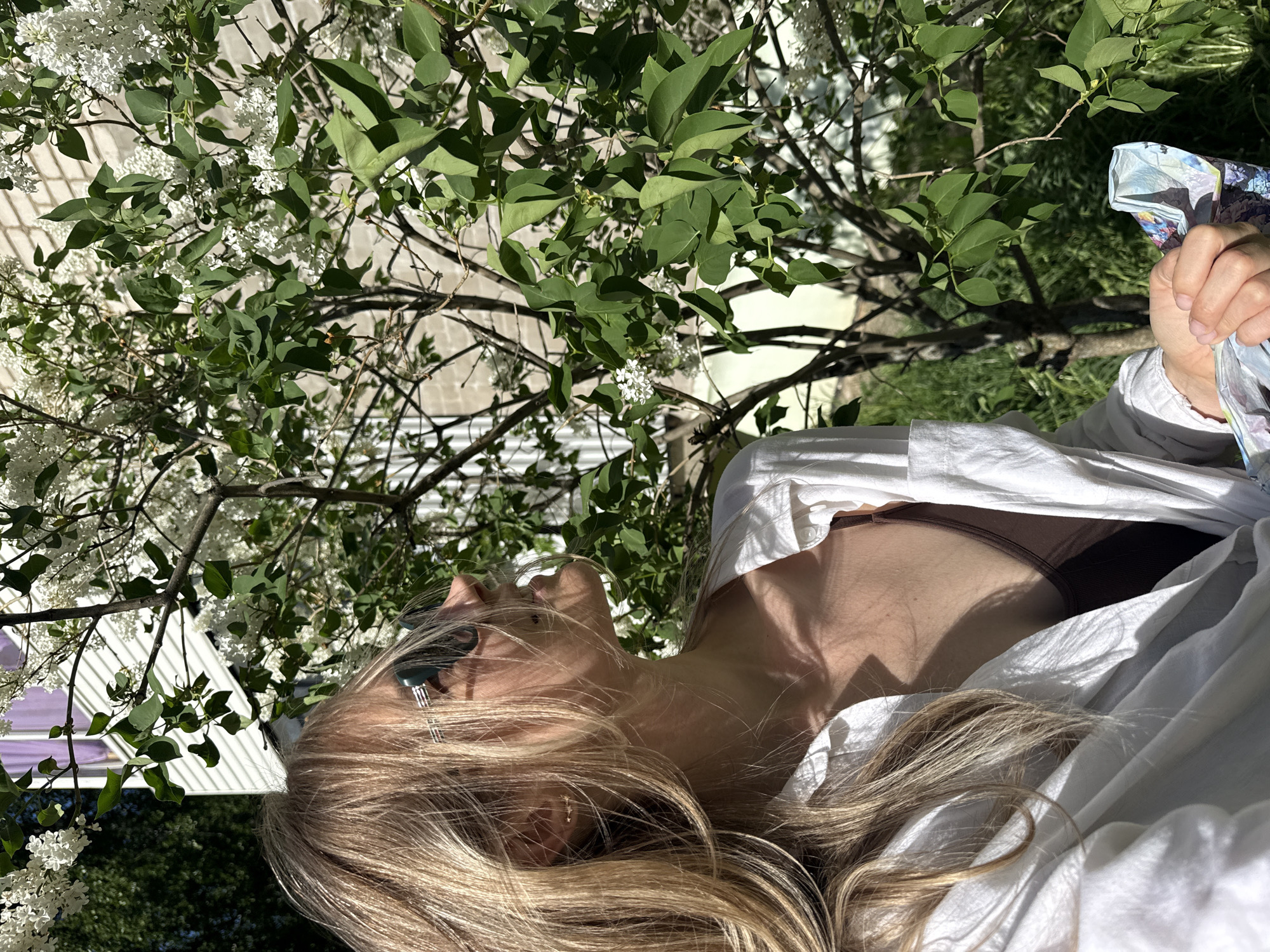 A woman lies under a tree with white flowers and green leaves. She wears sunglasses and a white shirt over a brown top. She is holding something in her right hand. The photo was taken outdoors, possibly in Chaikovsky, Russia.