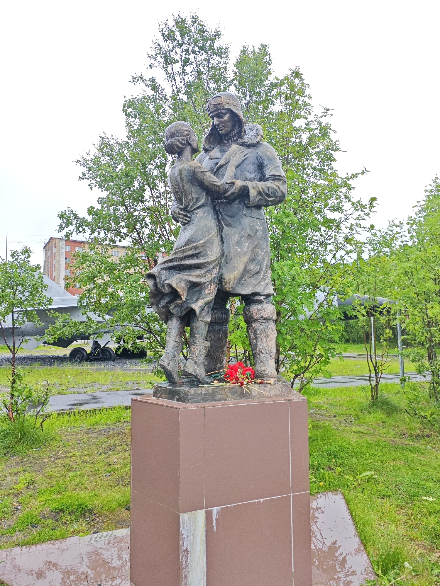 This media file features a bronze statue of a man and a woman embracing, situated in an outdoor setting. The man, dressed in what appears to be a pilot's uniform with a helmet and goggles, is holding the woman close. The woman is depicted in a flowing dress. Red flowers are placed at the base of the statue.
The statue stands on a reddish-brown pedestal in a grassy area. Behind the statue, trees with lush green foliage create a backdrop. In the distance, a multi-story building with red and orange brickwork is visible. To the left of the statue, part of an aircraft is visible, suggesting a possible connection to aviation history or a memorial site. A small yellow vehicle is parked near the aircraft. The ground around the statue is wet, indicating recent rain, and the sky appears overcast, suggesting it is a cloudy day.
The scene conveys a sense of warmth and connection between the two figures, possibly representing farewell, reunion, or enduring love amidst a challenging environment, given the context of Polyarny, Russia, which is a port city known for its naval aviation. The presence of the aircraft and the memorial-like arrangement of flowers further hint at a historical or commemorative context. No visible text is present in the image.