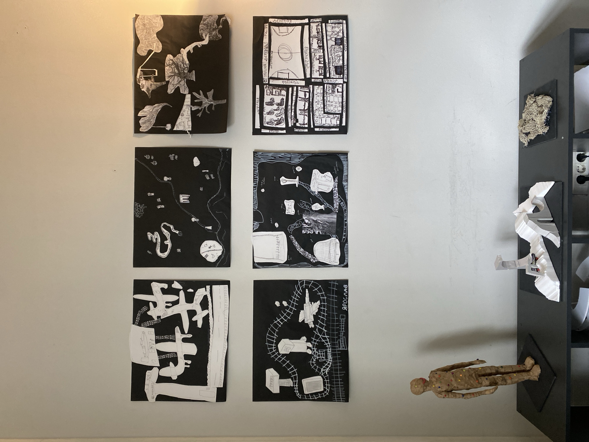 The image shows a wall with six black and white abstract drawings hanging on it. To the right of the drawings is a dark-colored shelf unit. The shelf unit contains a beige figure of a human lying flat, a white abstract sculpture, and a piece of dried plant matter. The wall is light colored. The location in Moscow, Russia is unknown.
