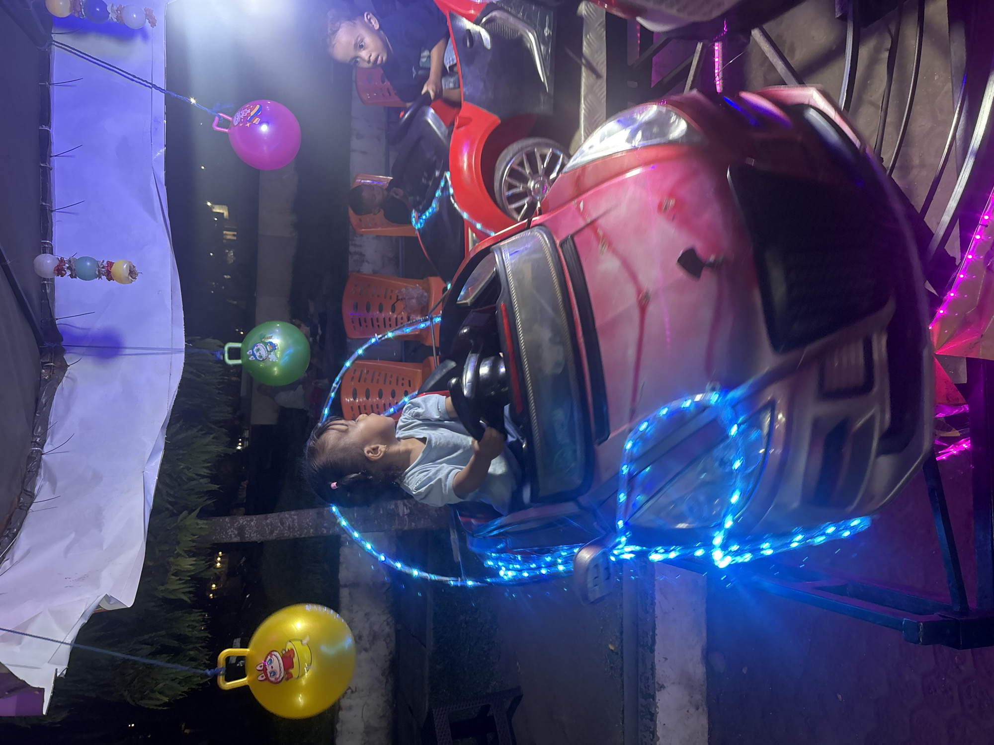 A young girl is riding a small pink toy car that is decorated with blue LED lights. She is holding onto the steering wheel and looking to her right. The car is on a ride at a night market in Lhokseumawe, Indonesia. In the background, other children are visible on similar rides. The ride is set up outdoors, under a canopy, with trees and buildings visible in the distance. There are also inflatable balls hanging from the ceiling, decorated with cartoon characters. The overall scene is festive and illuminated by colorful lights.