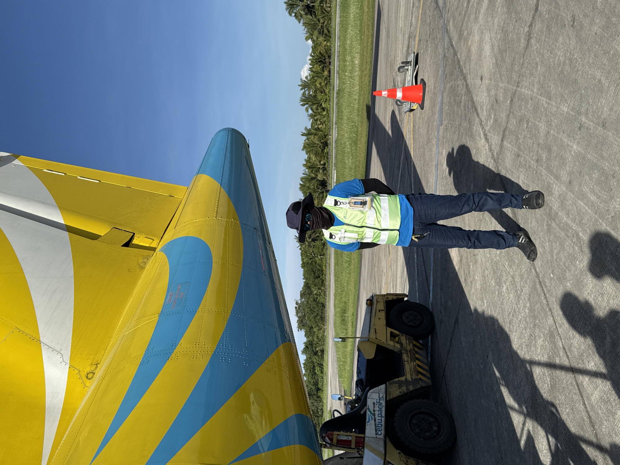 The image depicts an airport tarmac in Del Carmen, Philippines. A male airport worker is standing, wearing a bright green vest, dark pants, a blue shirt, a black face covering, sunglasses, and a hat. He is standing in front of a small yellow tow vehicle with black tires. Adjacent to him on the left is a section of an airplane, its tail painted in bright yellow, light blue, and white. Behind the worker is a grassy area, followed by a dense row of palm trees. A red and white safety cone is near the trees. The sky is clear and blue. The ground is concrete.