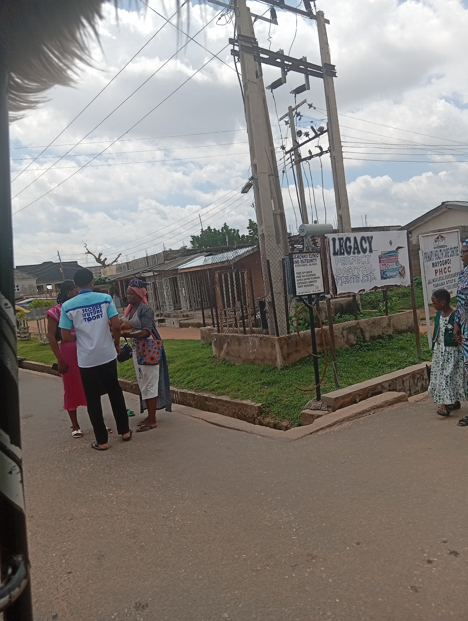 The image captures a street scene in Jalingo, Nigeria, under an overcast sky. In the foreground, a paved road is visible, with a sidewalk lined with grass and a low concrete barrier. On the sidewalk, three individuals are engaged in conversation. One person, wearing a light blue shirt with "SECURE FUTURE TODAY" printed on it, is facing away from the camera. Another person, wearing a pink dress, is partially visible to the left. A woman in a patterned headscarf and a long, dark grey dress is facing the other two. Behind them, several structures, including buildings and fences, are present. To the right, a large utility pole with multiple electrical wires and transformers dominates the background. A sign on the pole reads "LEGACY PERFECT DIGITAL PRINTS LTD." Another smaller sign next to it advertises "JA-KONKO CLINIC AND MATERNITY." Further to the right, another sign indicates "PRIMARY HEALTH CARE CENTRE MAYOGWOI PHCC." Two women and a child are walking on the sidewalk to the far right, with the child holding the hand of one of the women. The overall impression is of a busy, functional urban environment.