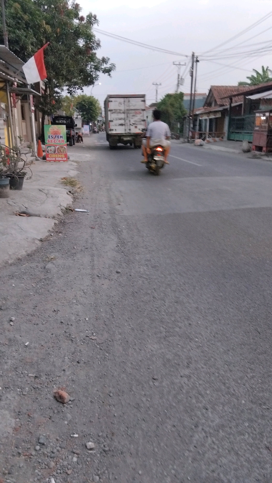 The image depicts a street scene in Tegal, Indonesia. In the foreground, a grey, unpaved shoulder borders a paved road. A small reddish-brown rock and scattered cigarette butts are visible on the unpaved shoulder. On the paved road, a motorcycle with two occupants is moving away from the camera. The rider wears a light-colored t-shirt and shorts, and the passenger appears to be wearing a darker shirt. Behind them, a white truck with a large cargo container is also traveling on the road. To the left of the road, a red and white Indonesian flag is affixed to a pole, and a sign advertising "ES TEH SEGAR" (Fresh Iced Tea) and "CIRENG KUAH SEBLAK" (Crispy Fried Snacks with Spicy Sauce) is propped up. The background shows buildings, trees, and utility poles under an overcast sky.