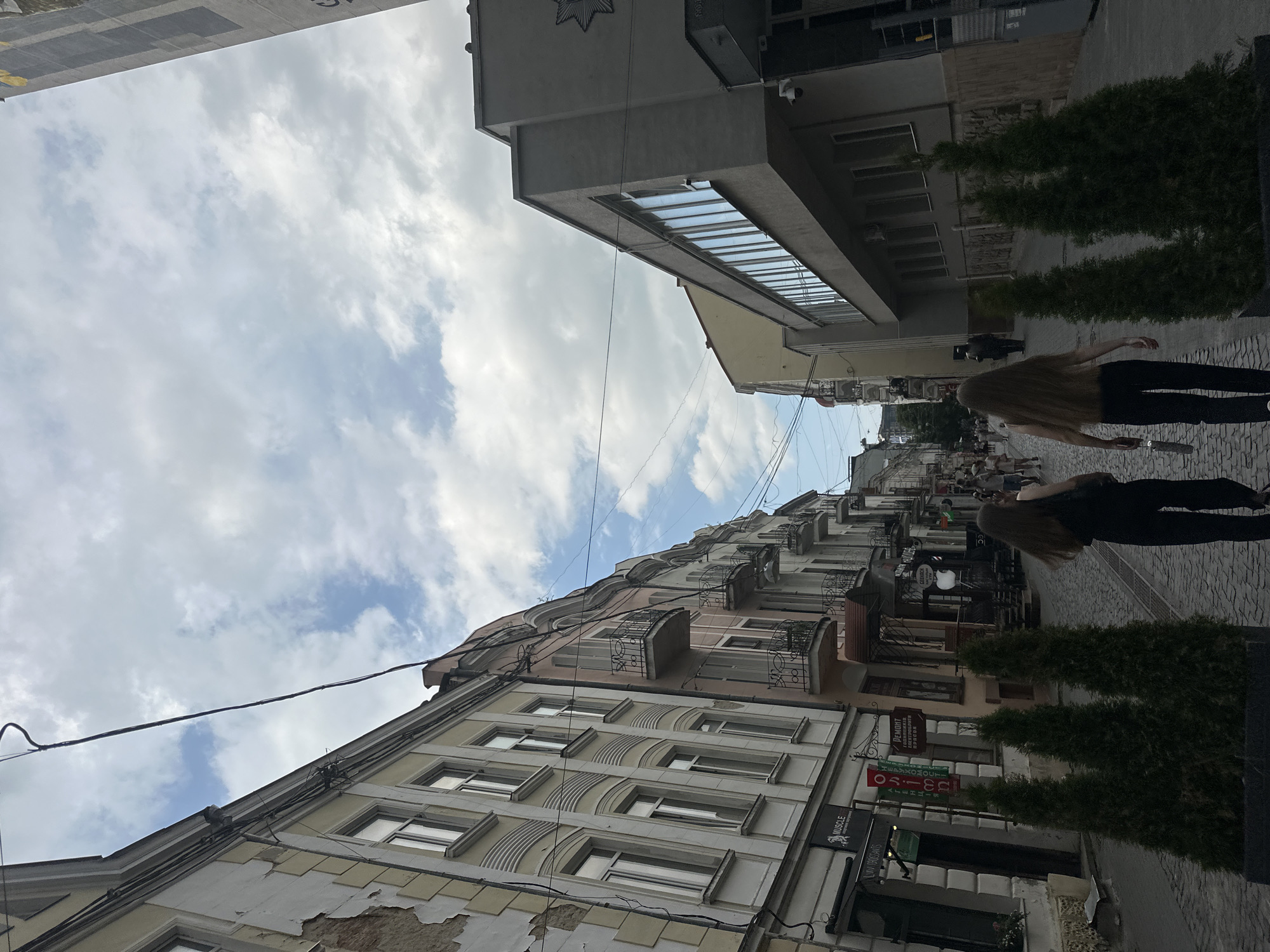 The image shows a street scene in Ternopil, Ukraine. Two young women, with long brown hair and wearing dark clothing, are walking away from the camera down a cobblestone street. They are walking on the right side of the street, on the sidewalk. The street is lined with buildings on both sides. On the left, there are older buildings with ornate facades and balconies. On the right, there is a more modern, grey building with a long row of windows. Several evergreen trees in planters are positioned along the sidewalk. In the background, more people are visible walking along the street, and there are various shop signs on the buildings. The sky is partly cloudy.