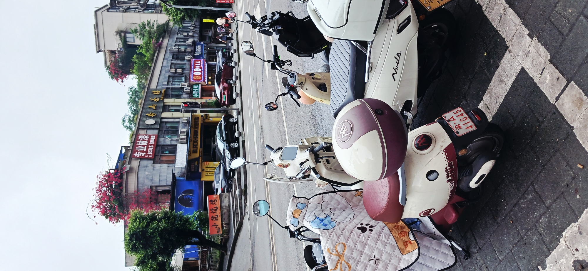 The image depicts a street scene in Jiulongpo District, China, with parked scooters dominating the foreground. Several scooters are lined up in a row, some with protective seat covers and helmets. Buildings are visible across the street, featuring commercial signs in Chinese characters, including the names of businesses such as "小院烧烤", "BLACKHAWK", "小程生鲜", and "专业做漆", indicating various commercial establishments. Cars are parked along the street. The image captures a typical urban environment with a mix of vehicles and commercial activities. A license plate is visible, reading "渝・A41645". The scooters are parked on a paved surface with white painted lines. There are trees and bougainvillea flowers adding a touch of greenery to the scene. The writing "NSLA" is visible on one of the scooters.