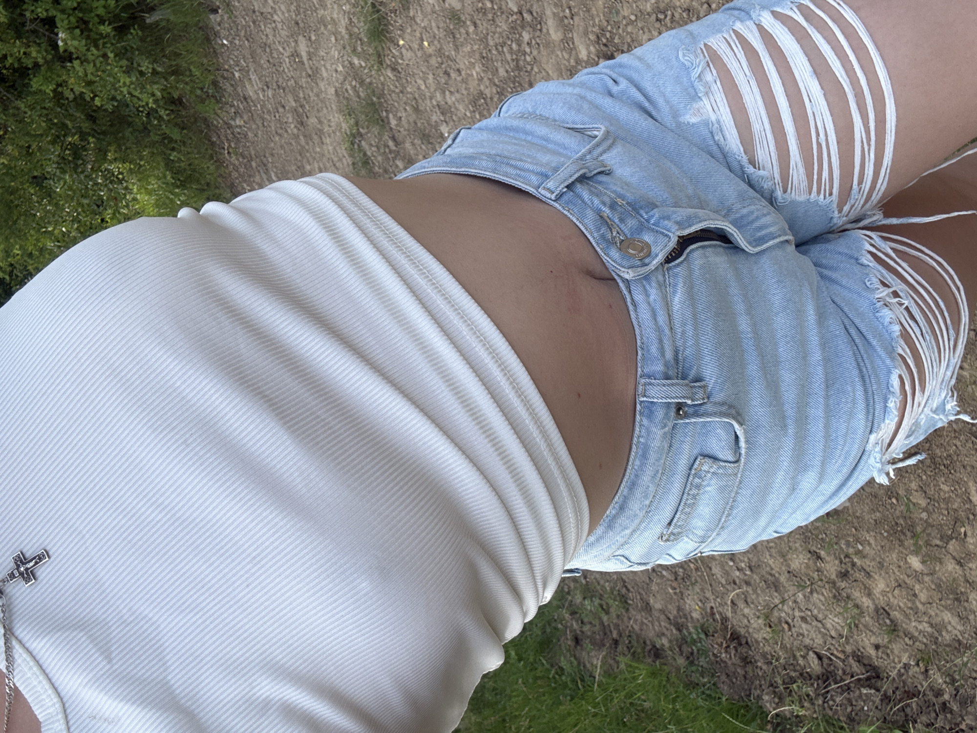 A person is standing outdoors, on a dirt path with grass and trees in the background. They are wearing a white ribbed crop top and light-wash denim shorts with rips and frayed edges. A silver cross necklace is visible on the person's chest. The lighting suggests it is daytime. The location is identified as Ilnytsia, Ukraine.