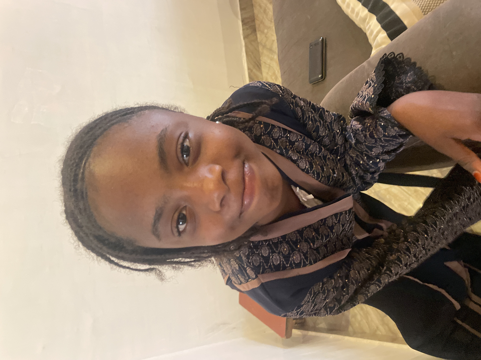 A young woman with braided hair is reclining on a gray upholstered couch. She is wearing a navy blue garment with brown trim and embellished with a patterned, glittering design. A dark cell phone rests on the couch beside her. The background includes a beige wall and striped cushions on the couch. The location, Jos, Nigeria, cannot be verified from the image.