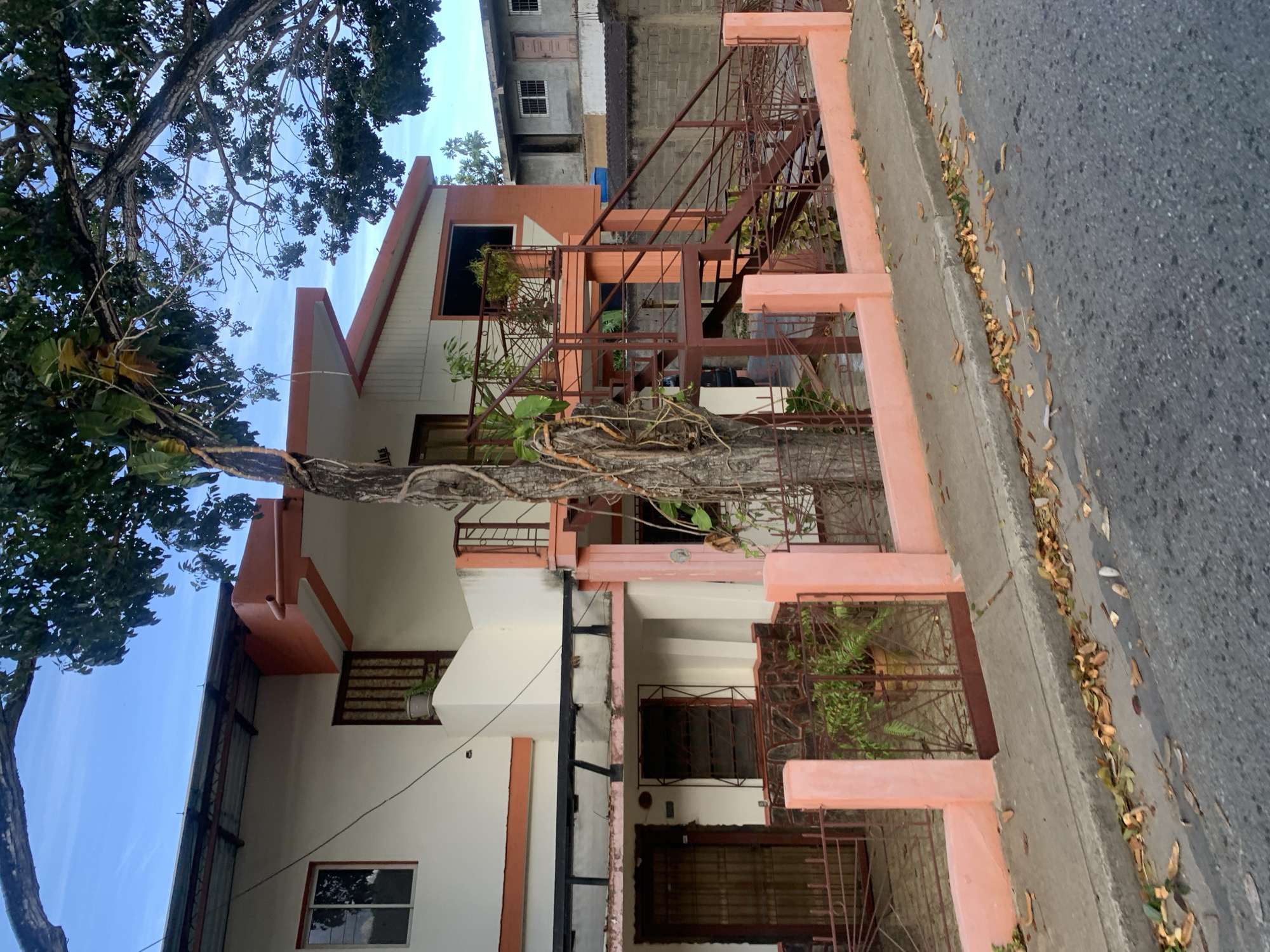 This image depicts the exterior of a two-story residential building located on a street in La Asuncion, Venezuela. The building is painted white with orange trim and features an external metal staircase leading to the upper level. A large tree with visible vines grows in front of the building, partially obscuring the staircase and the building's facade. A pink-colored fence with decorative metalwork encloses the property. The street in front of the building is paved, with a concrete sidewalk. Fallen leaves are scattered on the sidewalk and the street. In the background, another residential building with a gray facade and barred windows can be seen, along with a concrete block wall. The sky is visible at the top of the image, with some clouds. There are no people visible in the image.