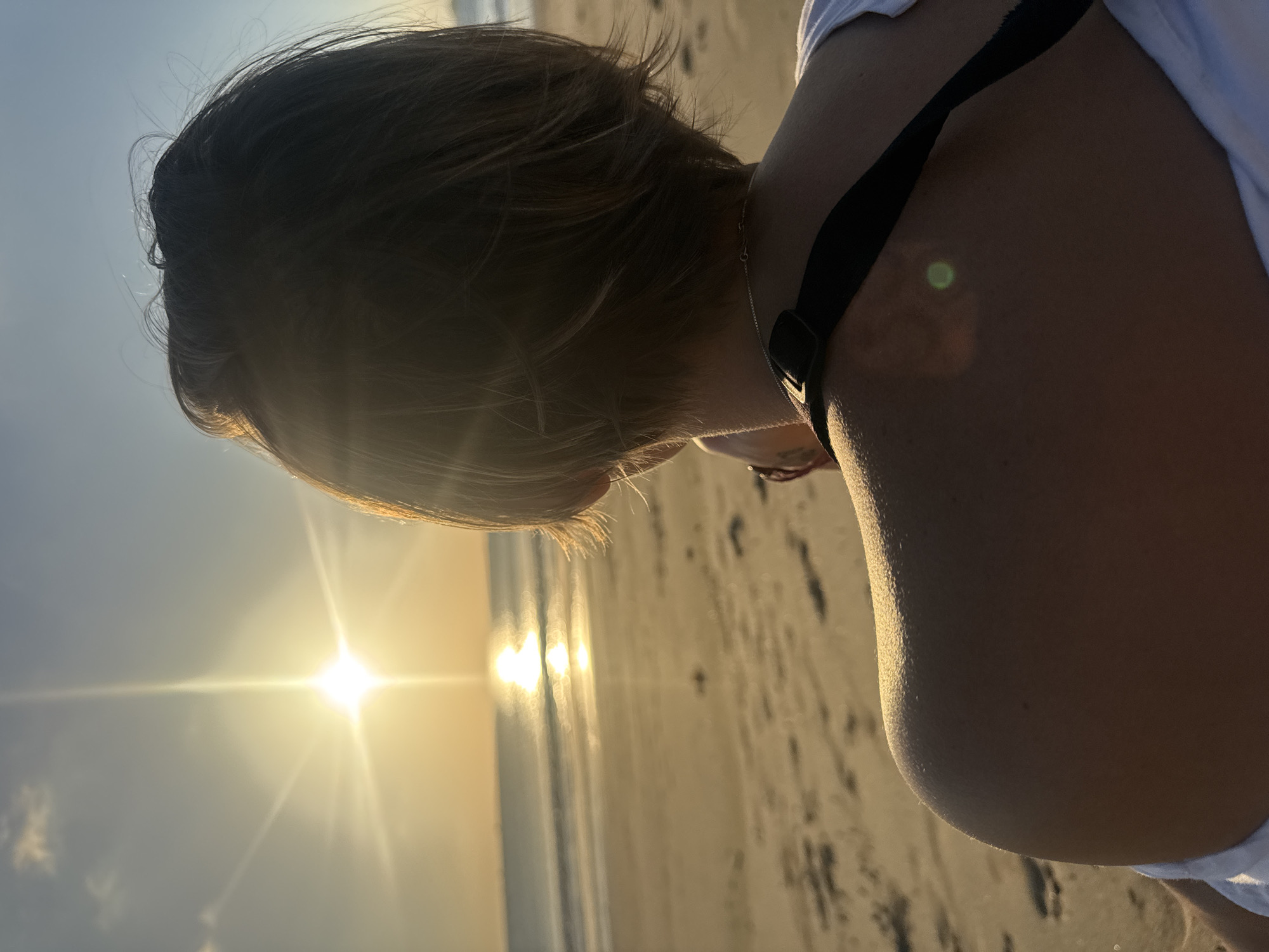 A person seen from behind stands on a beach in Kuta, Indonesia, facing the ocean at sunset. The sun is low on the horizon, casting a bright glare that reflects on the water and creates a starburst effect in the sky. The person has short, light brown hair and is wearing a white shirt. Their left shoulder and upper back are visible, illuminated by the sun. The sand of the beach is visible in the foreground, with some footprints. The ocean is calm, with gentle waves. The sky is clear with a slight gradient of blue and orange.