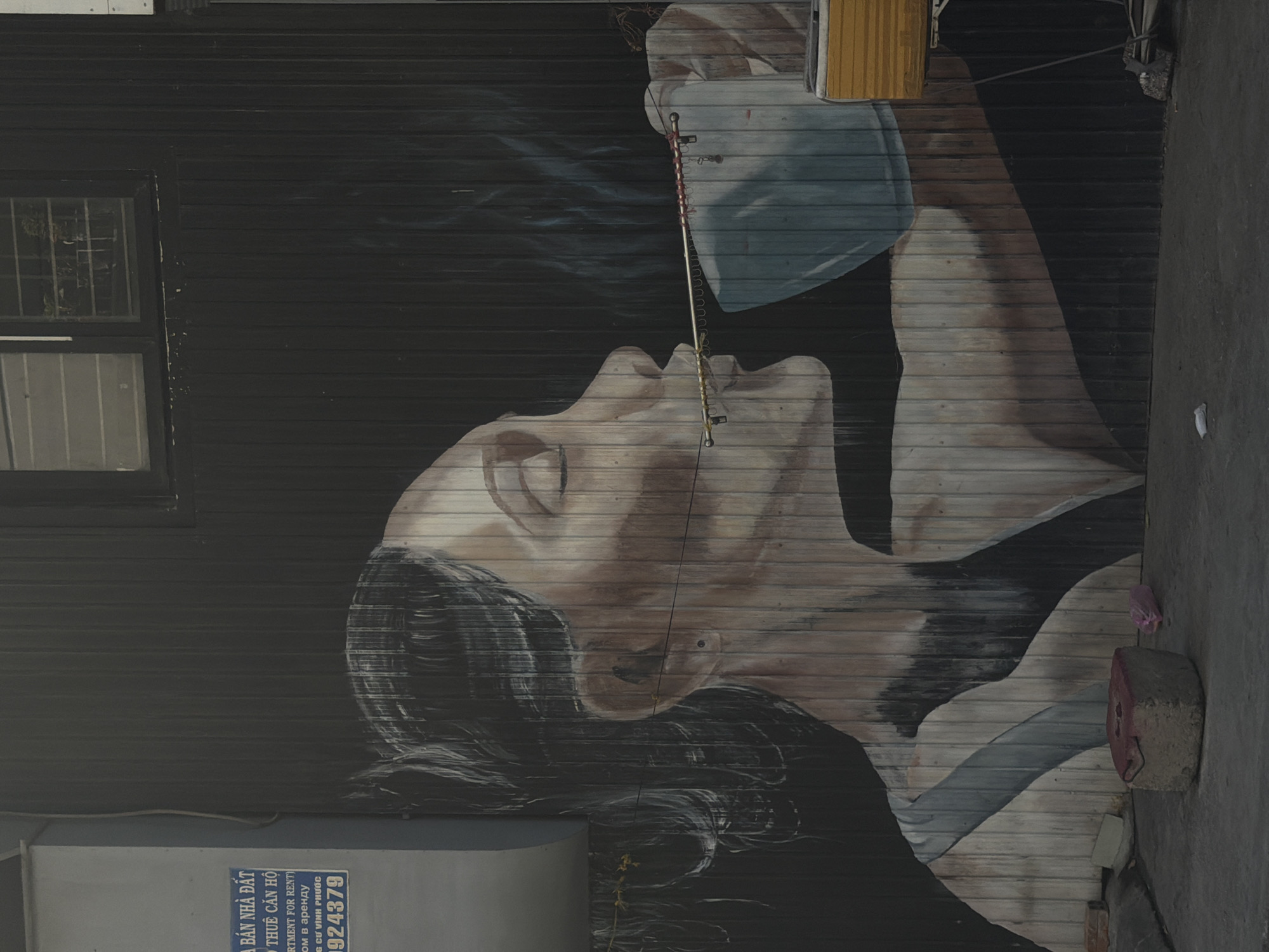 A mural of a woman's profile, appearing to drink from a light blue mug, is painted on a corrugated metal wall in Bac Nha Trang Ward, Vietnam. The woman has her eyes closed and dark hair. A thin metal rod, adorned with red string and what appear to be charms or keys, extends from her mouth to the mug. The mug is held by an unseen hand or is part of the mural's composition. Smoke or steam rises from the mug. To the left of the mural, a light grey structure with blue signage is visible. The signage includes Vietnamese text and the number "9924379". In the foreground, on the ground, are several objects including a square concrete block with a reddish top, a pink plastic bag, and a white piece of paper. The ground appears to be concrete. The overall environment suggests an urban or semi-urban outdoor setting.