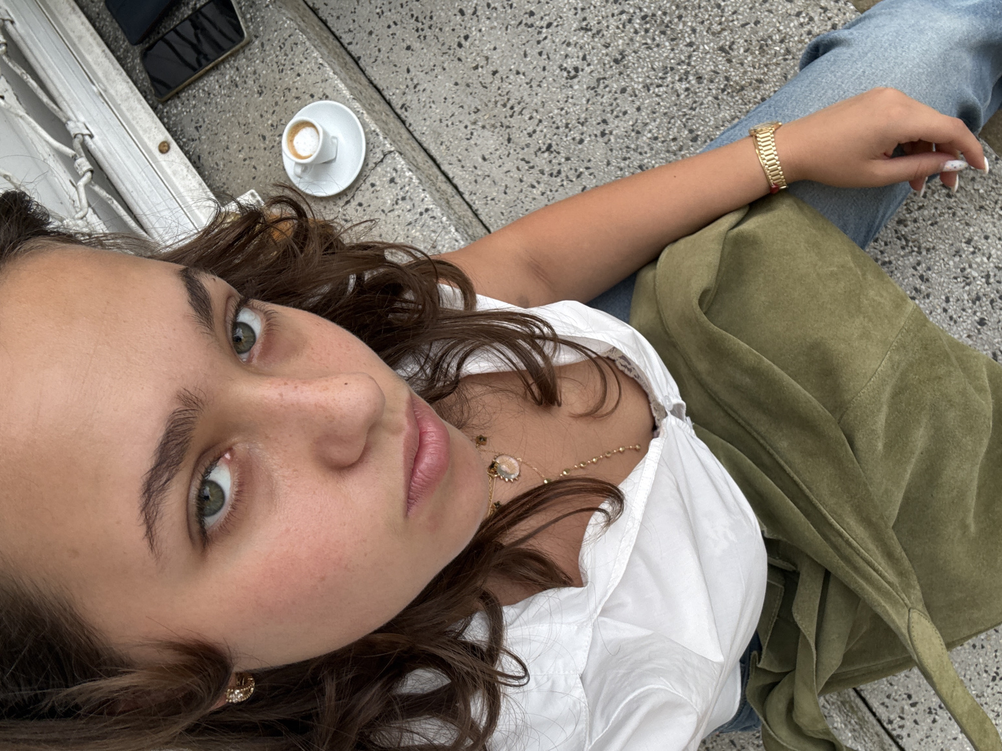 The image features a close-up, overhead shot of a young woman lying on a textured, gray, concrete surface. She is wearing a white, collared shirt and blue jeans. A green, suede jacket is draped over her lap. She is also wearing a gold bracelet on her left wrist and a delicate gold necklace with a pendant. Her expression is relaxed, with her eyes looking up and slightly to the left, revealing green irises. Her hair is dark brown and wavy, framing her face. In the upper left corner of the frame, a small white cup of coffee sits on a saucer, and a dark-colored smartphone is visible. The scene appears to be outdoors, given the lighting and the concrete ground, possibly a cafe or a public seating area. The time of day is likely daytime, suggested by the bright, natural light illuminating the scene. There is no visible text in the image.