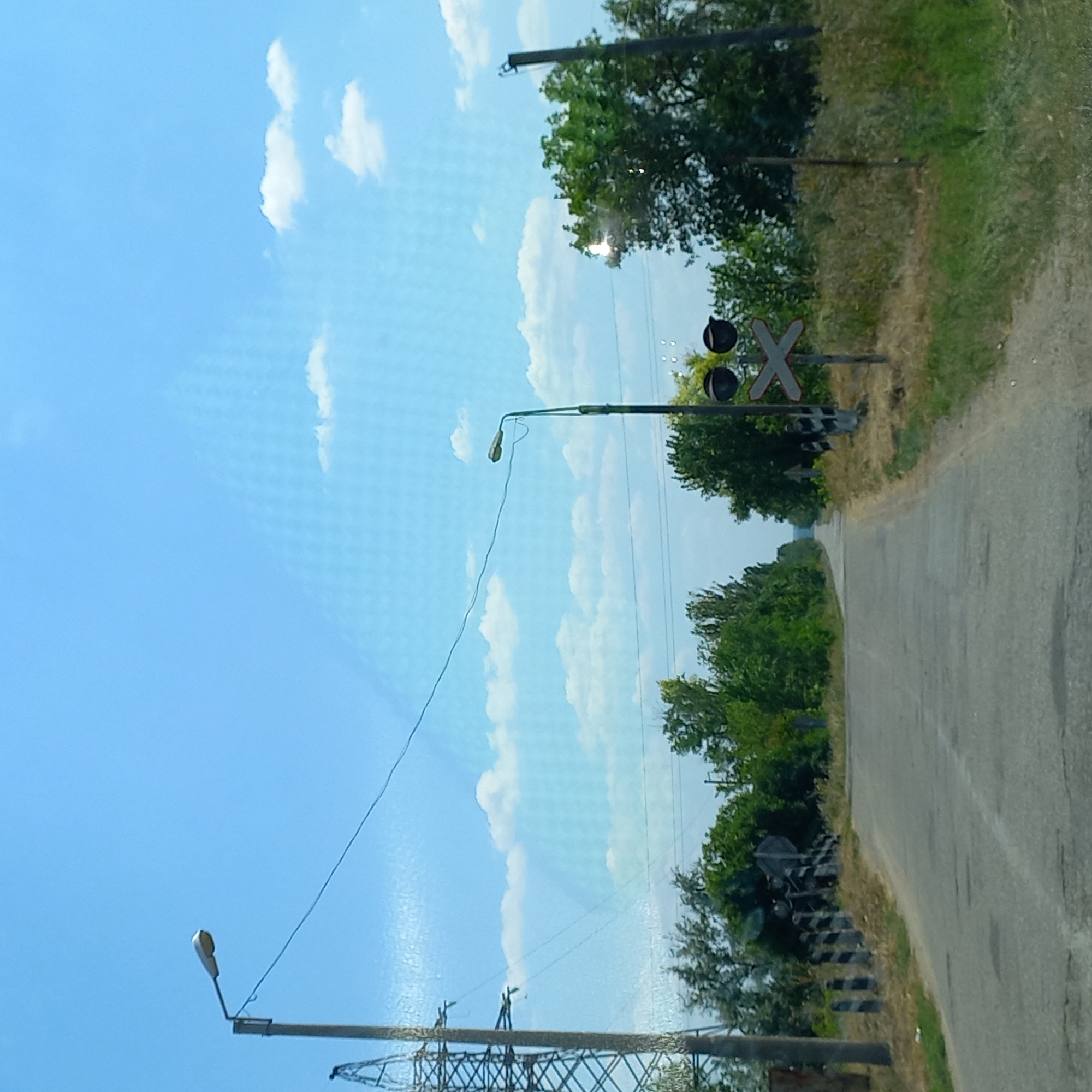 The image depicts a rural road with a clear blue sky and scattered white clouds. On the left side of the road, a tall power line tower is visible, with a lamppost on a pole extending over the road. Further down the road, another lamppost is positioned on the right side. A railroad crossing sign, featuring a white 'X' with two black circles above it, is also present on the right. The road is asphalt and appears to be in moderate condition, with some cracks and patches. The surrounding environment is characterized by dry, grassy fields and dense green trees. The scene is devoid of any people or vehicles. The presence of the power line tower and the railroad crossing suggests this location is on the outskirts of a populated area, possibly Klyntsi, Ukraine, given the context provided.