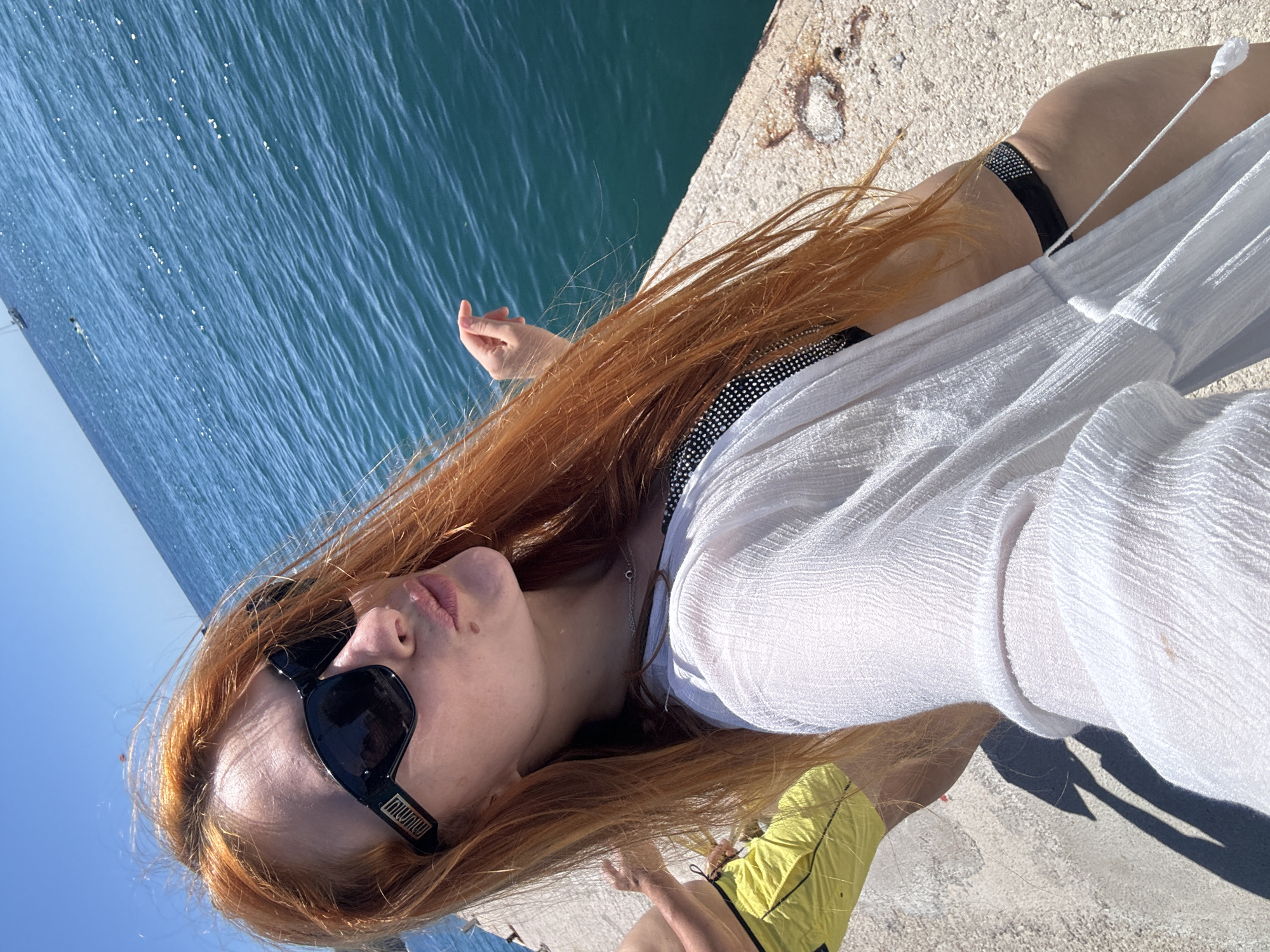 An overhead shot captures a young woman relaxing on a concrete pier overlooking clear blue water in Budva, Montenegro, under a bright, sunny sky. The image is tilted diagonally, giving a dynamic perspective. The woman has long, flowing reddish-blonde hair and is wearing black rectangular sunglasses with "MIU MIU" visible on the arm. She has a subtle mole near her lips, which are slightly parted.
She is dressed in a sheer white cover-up top over a black top adorned with sparkling embellishments, likely a bikini top, and yellow shorts. A delicate silver necklace with a small charm hangs around her neck. Her left hand is visible near the water's edge, fingers slightly curled, while her right arm and legs are partially visible towards the bottom right.
The backdrop features an expansive body of blue water, shimmering with sunlight and showing gentle ripples. The concrete pier she rests on is light-colored and somewhat weathered. Strong shadows indicate it is daytime, likely mid-morning or afternoon, with clear weather. The overall impression is one of leisure and enjoyment of a sunny day by the sea.