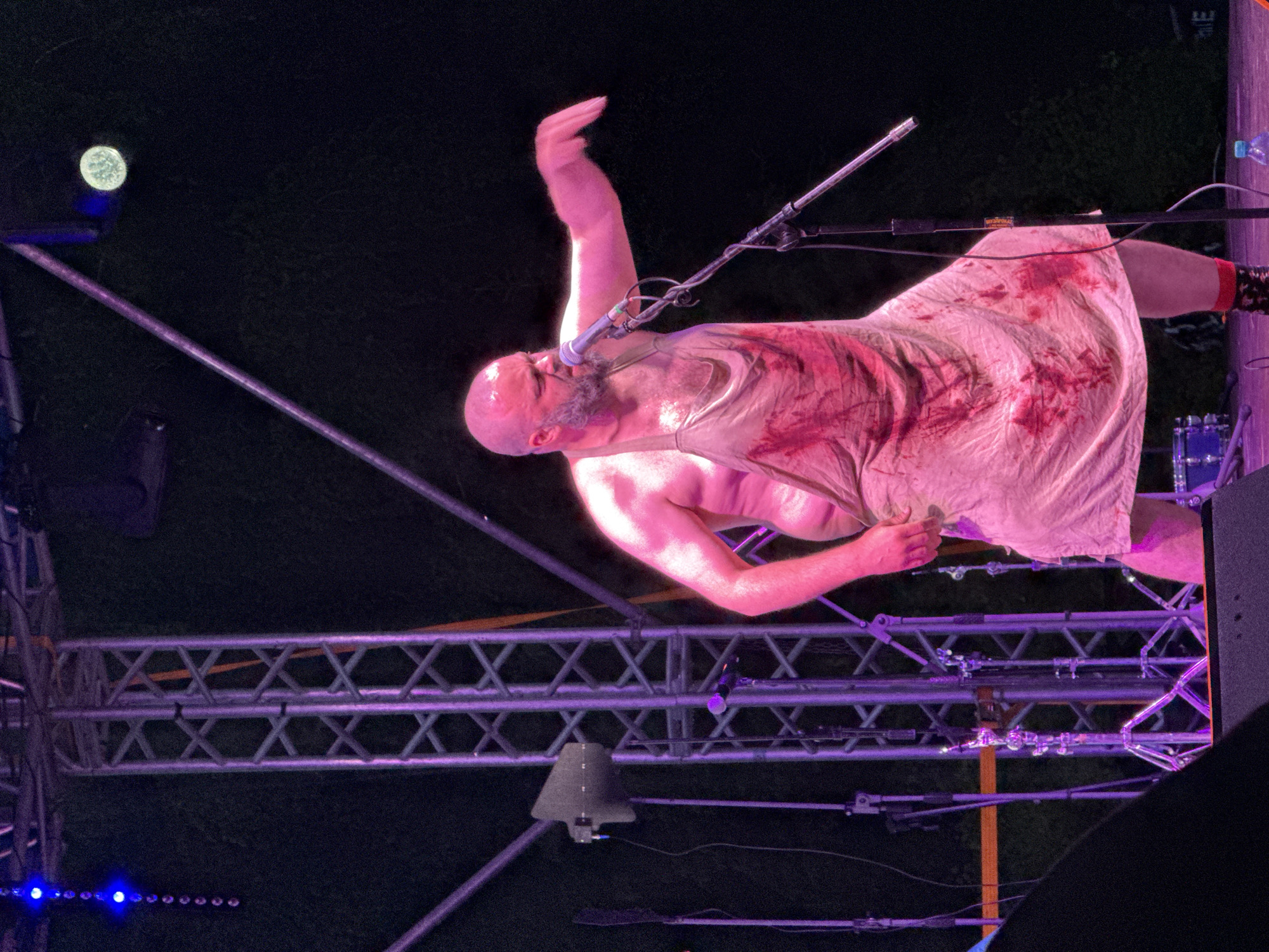 The image depicts a bald, bearded man, appearing to be in his late 30s to 50s with a robust build, performing on an outdoor stage at night in Большая Кивара, Russia. He is bare-chested and wears a light-colored, possibly off-white, sleeveless apron heavily stained with multiple patches of dark red liquid, consistent with simulated blood.
The man's head is bald, and he has a full, greyish beard. His facial expression is intense, with his mouth open, indicating he is singing or shouting into a silver-colored microphone mounted on a black stand positioned directly in front of him. His left arm is extended outwards, bent at the elbow, with fingers slightly curled, while his right arm is bent at the elbow, with his hand near his side. His right foot, clad in a red and black patterned sock, is visible on the stage floor.
The stage features prominent silver-colored metal truss structures with cross-bracing, illuminated by purple stage lighting. In the upper left background, a vertical string of small blue lights is visible. A circular, light-colored spotlight is present in the upper right background. Additional stage equipment includes a dark grey wedge-shaped monitor speaker on the left side, multiple black microphone stands, and a partial view of a blue drum from a drum kit on the lower left. Various black cables run across the stage floor. A clear plastic water bottle with a blue cap rests on the stage floor in the lower right corner. The background beyond the stage structures is predominantly dark, suggesting nighttime, with subtle hints of dark green foliage visible in places.