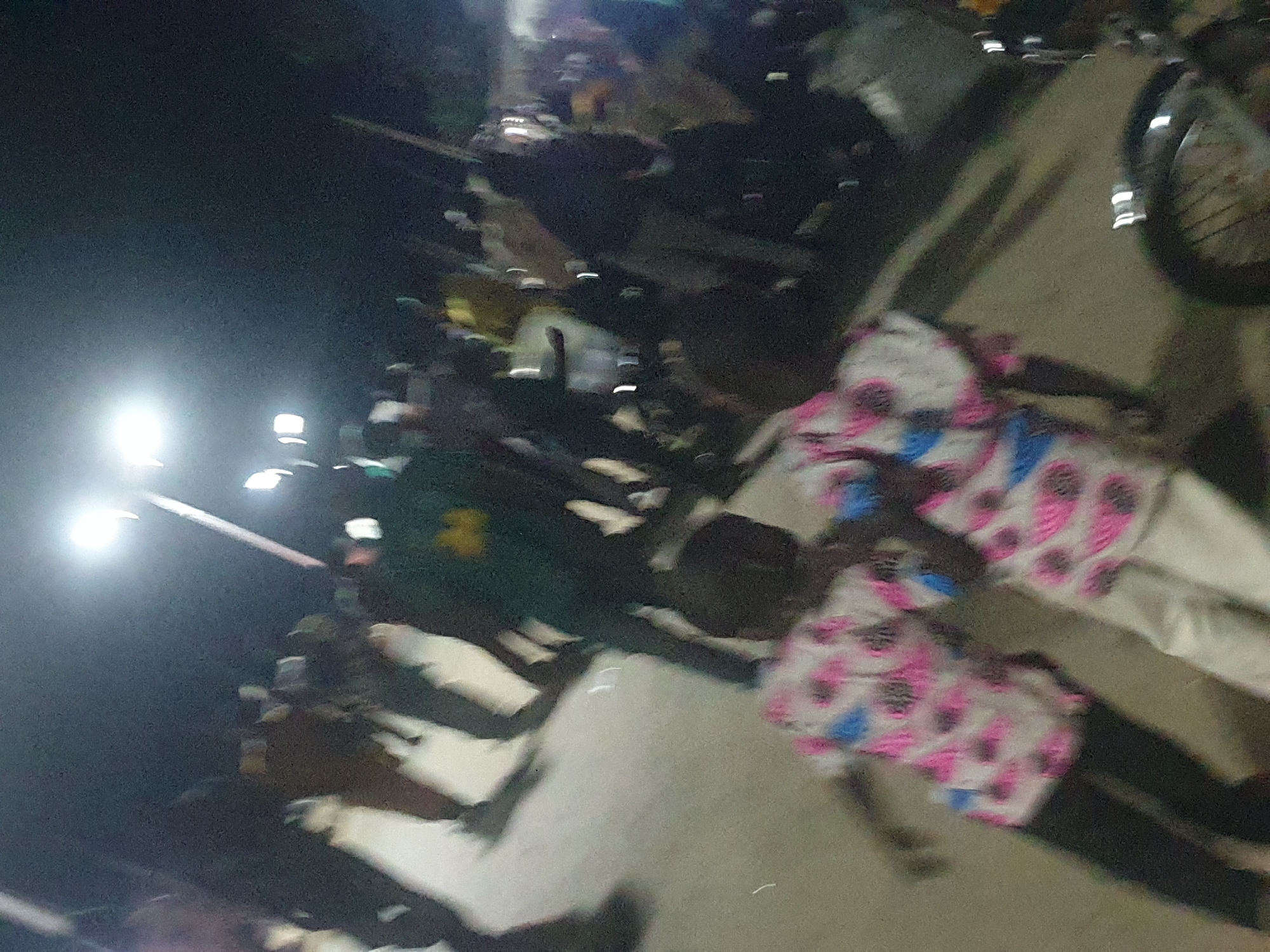 The image shows a street scene at night in Zango-ndaji, Nigeria. A group of people are gathered, illuminated by streetlights. A bicycle wheel is visible on the edge of the frame. Some individuals are lying on the ground. One person is wearing a shirt with a pink and blue pattern. Another is wearing a green shirt.